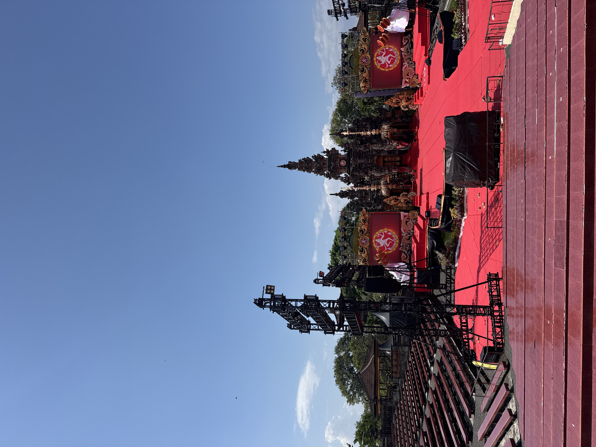 The image shows an outdoor event setup in Denpasar, Indonesia. A vibrant red carpet stretches across a large, open area, flanked by rows of red tiered seating. In the background, elaborate stage structures and scaffolding are visible, suggesting a performance or festival. Two large red banners with circular emblems featuring a triquetra symbol and a stylized figure are prominently displayed on the stage. The scene is set against a clear blue sky with scattered white clouds, indicating fair weather during the daytime. Lush green trees and foliage are visible around the stage area. The overall impression is one of a prepared outdoor venue for a significant cultural or entertainment event.
