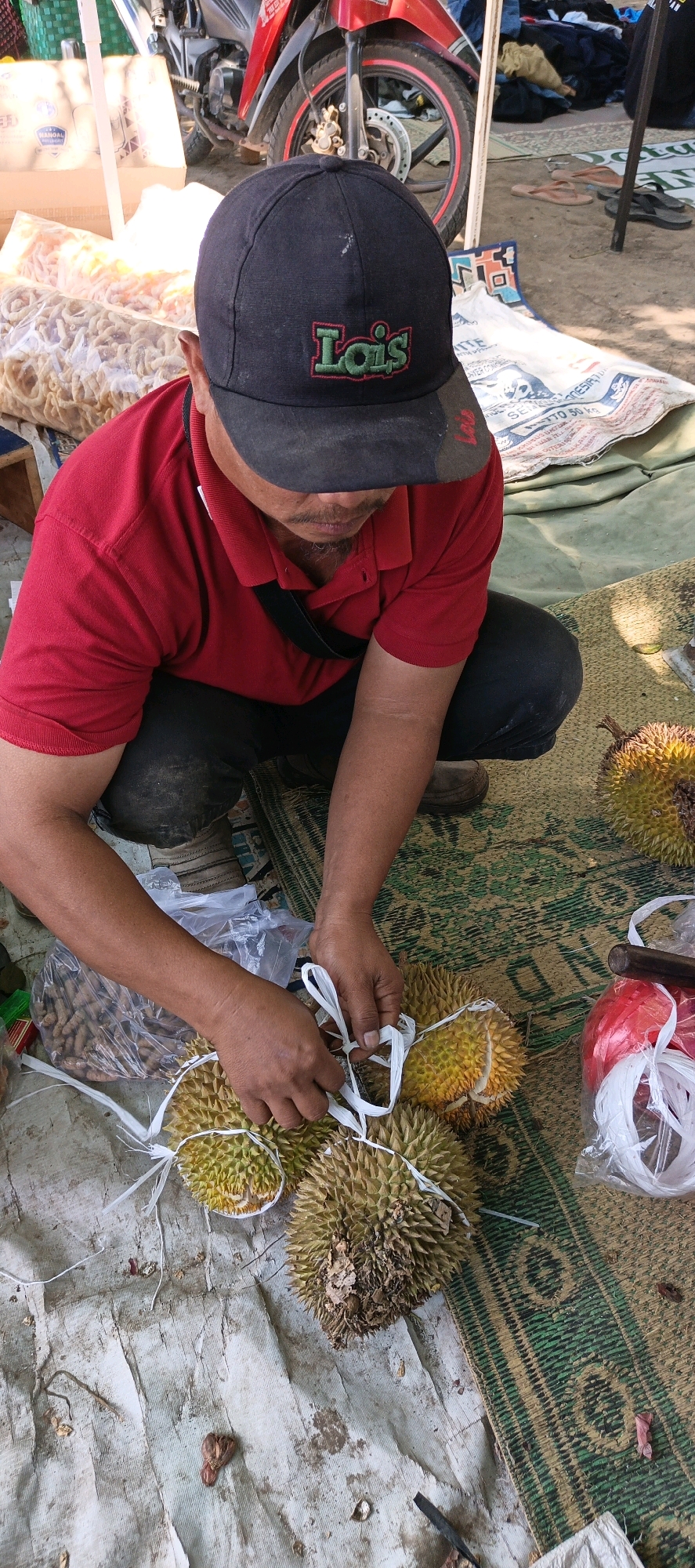 An overhead medium shot captures a man in Jatinom, Indonesia, squatting on a woven mat next to a collection of durians he is preparing. The man, wearing a red polo shirt and a black baseball cap with the word "Lois" embroidered in green and red, is in the process of tying several durians together with white string. His hands are actively engaged in the task.
The durians are the central focus, their spiky green and yellow exteriors visible. Some appear ripe, with portions of their flesh exposed. Beside the durians, there are bags of what appear to be snacks or nuts, and a red plastic container filled with white string.
The environment suggests an outdoor market setting. Behind the man, a red motorcycle is partially visible, along with what looks like merchandise displayed on tables or racks. The ground beneath the mat is sandy or dirt. The overall scene conveys a moment of commerce and food preparation in a bustling local market.