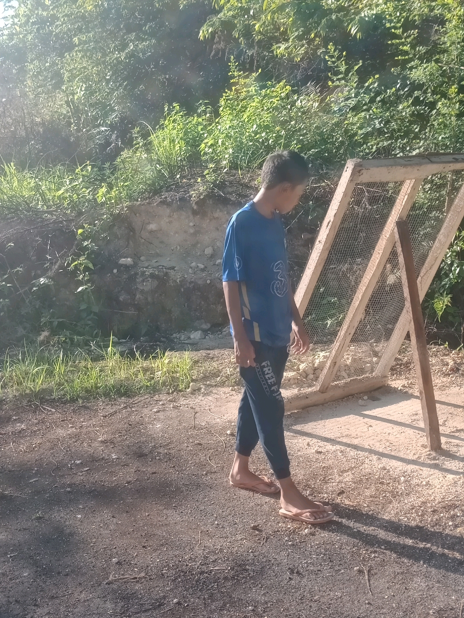 A young male, appearing to be of Indonesian ethnicity, is depicted in the foreground of an outdoor scene. He is wearing a blue t-shirt with a logo and dark-colored pants with a striped pattern. He is also wearing flip-flops. The subject is walking on a dirt path, with a partially constructed wooden frame covered in mesh behind him to his right. The environment is lush with green vegetation in the background, suggesting a rural or semi-rural location. The lighting indicates it is daytime, possibly late afternoon given the long shadows. The scene is consistent with a location in Cintakarya, Indonesia, characterized by natural landscapes and outdoor construction or agricultural activities.