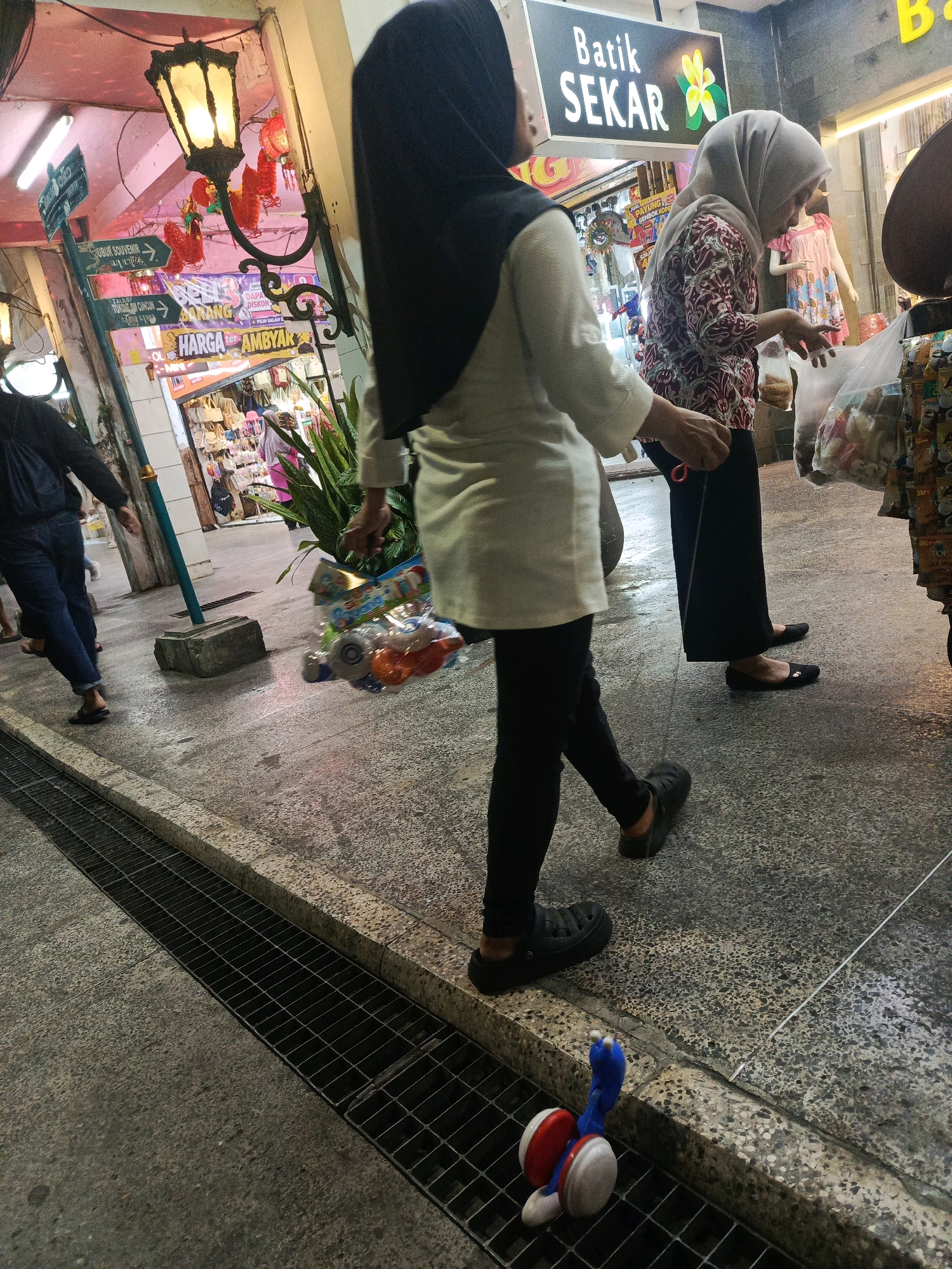 The image captures a bustling street scene in Yogyakarta, Indonesia, likely in an area with shops and markets. In the foreground, a person wearing a black hijab and white tunic walks from right to left, carrying a plastic bag filled with what appear to be toys. To the right, another person, also wearing a hijab and a patterned blouse, is seen from the waist down, holding a clear plastic bag with items. Behind them, a shop displaying various goods is visible, with signs in Indonesian. The street is paved with textured concrete, and a drainage grate runs along the edge of the walkway. In the background, a traditional lamppost with ornate detailing illuminates the scene, casting a warm glow. Red lanterns are also visible, adding to the festive atmosphere. The overall impression is one of a vibrant commercial district in Yogyakarta.