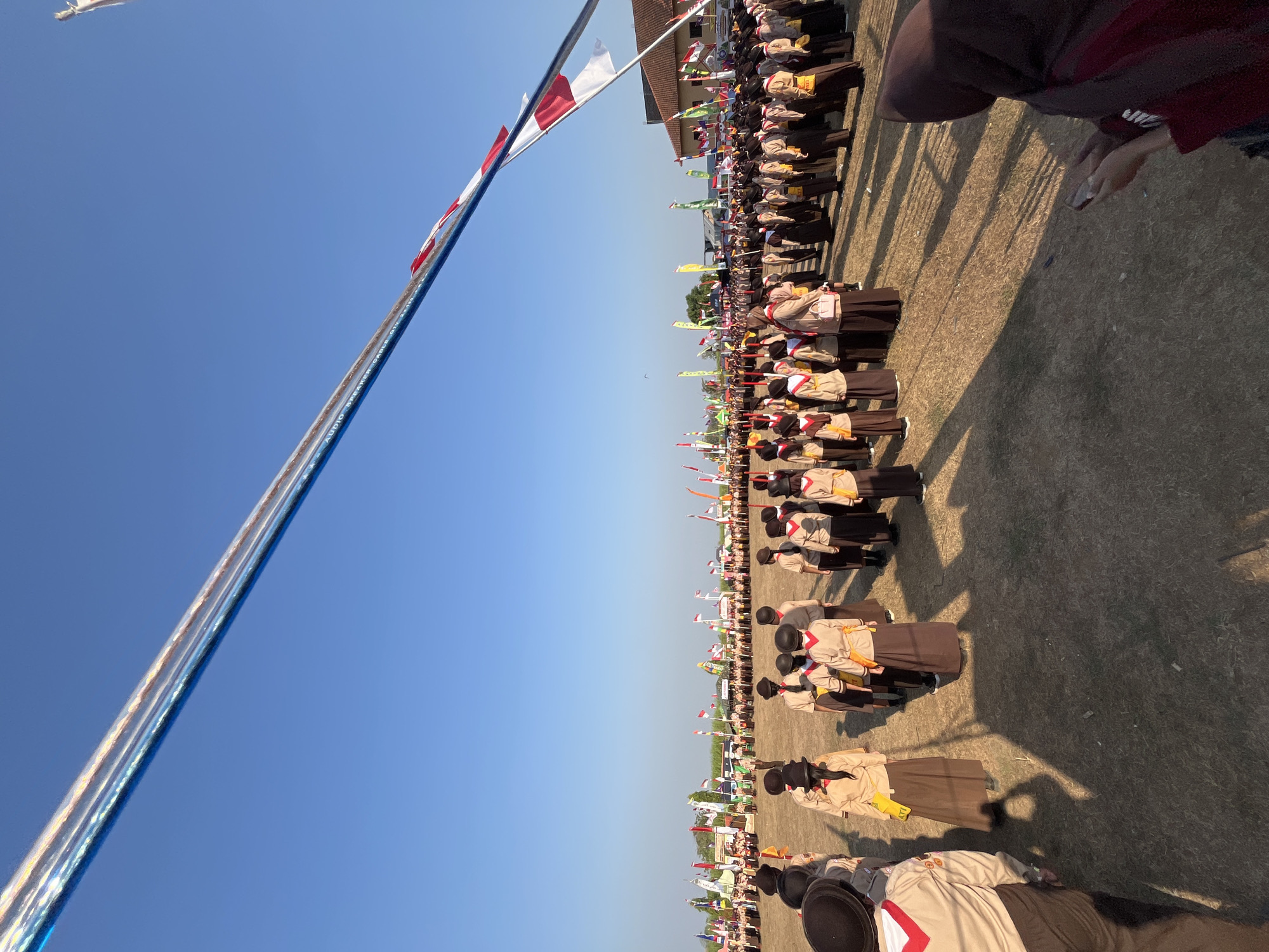 In Gembong, Indonesia, a large group of scouts in uniform stand in formation on a dry, grassy field under a clear blue sky. The scouts, mostly young individuals, wear tan shirts with brown skirts or pants and distinctive brown hats. Many have red and white neckerchiefs. In the background, a line of various flags, including Indonesian flags, are visible. The sun casts long shadows across the field, indicating it is either early morning or late afternoon. A building with a tiled roof is visible in the far background. The scene appears to be an outdoor event or ceremony.