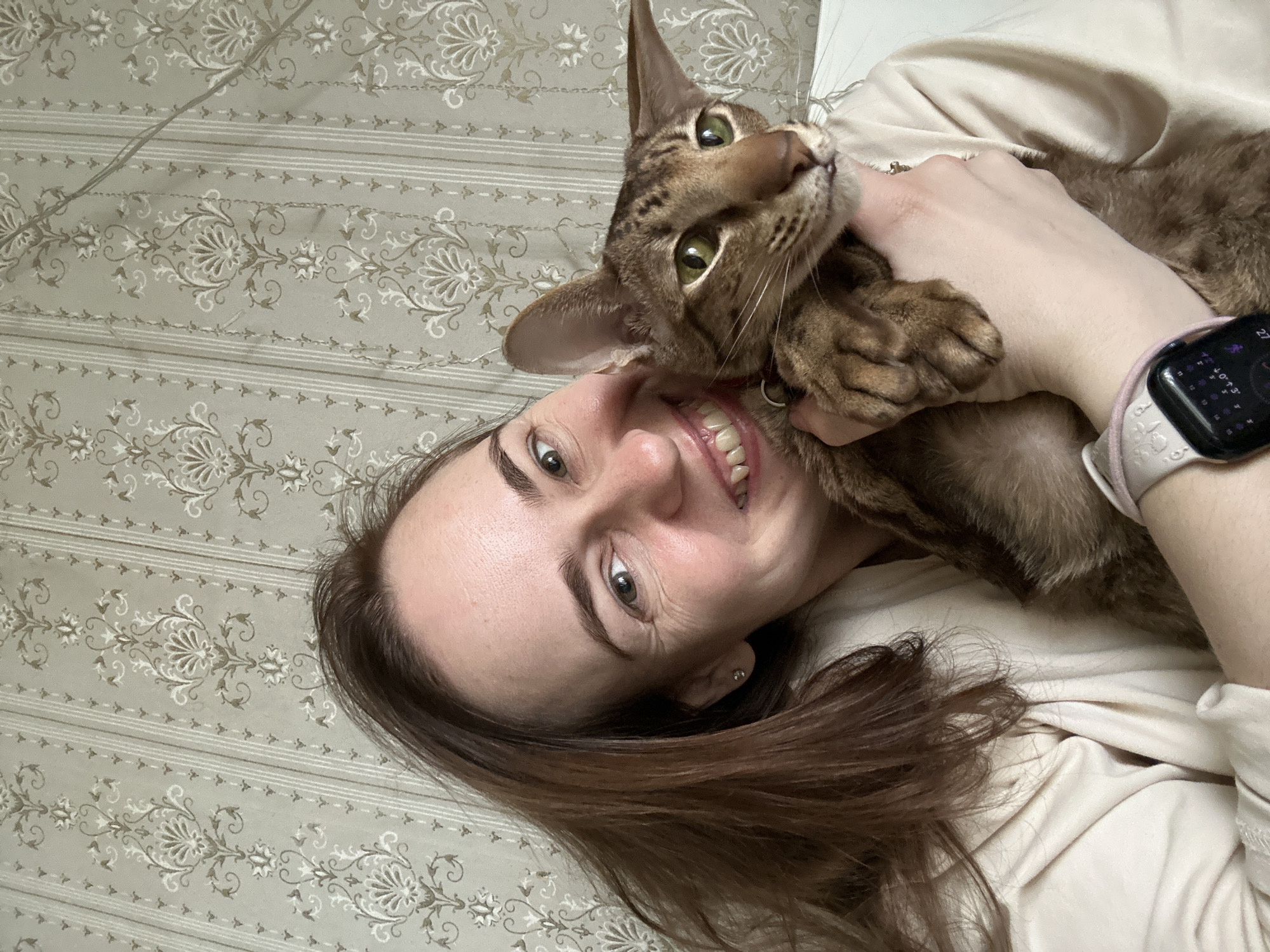 An overhead, close-up shot captures a joyful woman embracing her cat in an indoor setting. The woman, with brown hair and light skin, is lying down and smiling broadly at the camera, her teeth visible. She has green eyes and wears a small stud earring in her right ear. A smartwatch with a light pink band is on her left wrist, displaying what appears to be activity rings and numbers, including "27" and "0->3."
She is holding a short-haired, brown tabby cat with prominent ears and striking green eyes. The cat is also looking towards the camera with an alert, curious expression, its head slightly tilted. Its front paws are visible, held near the woman's hand. The woman's light-colored top is visible around her arm and shoulder.
The background consists of a patterned surface, likely wallpaper or bedding, in a light grey or beige hue, decorated with intricate white and darker beige floral and scroll designs, along with horizontal striped patterns. The lighting is soft and even. The image conveys a warm, affectionate moment between a pet owner and their cat. The location context provided for the image is Moscow, Russia.