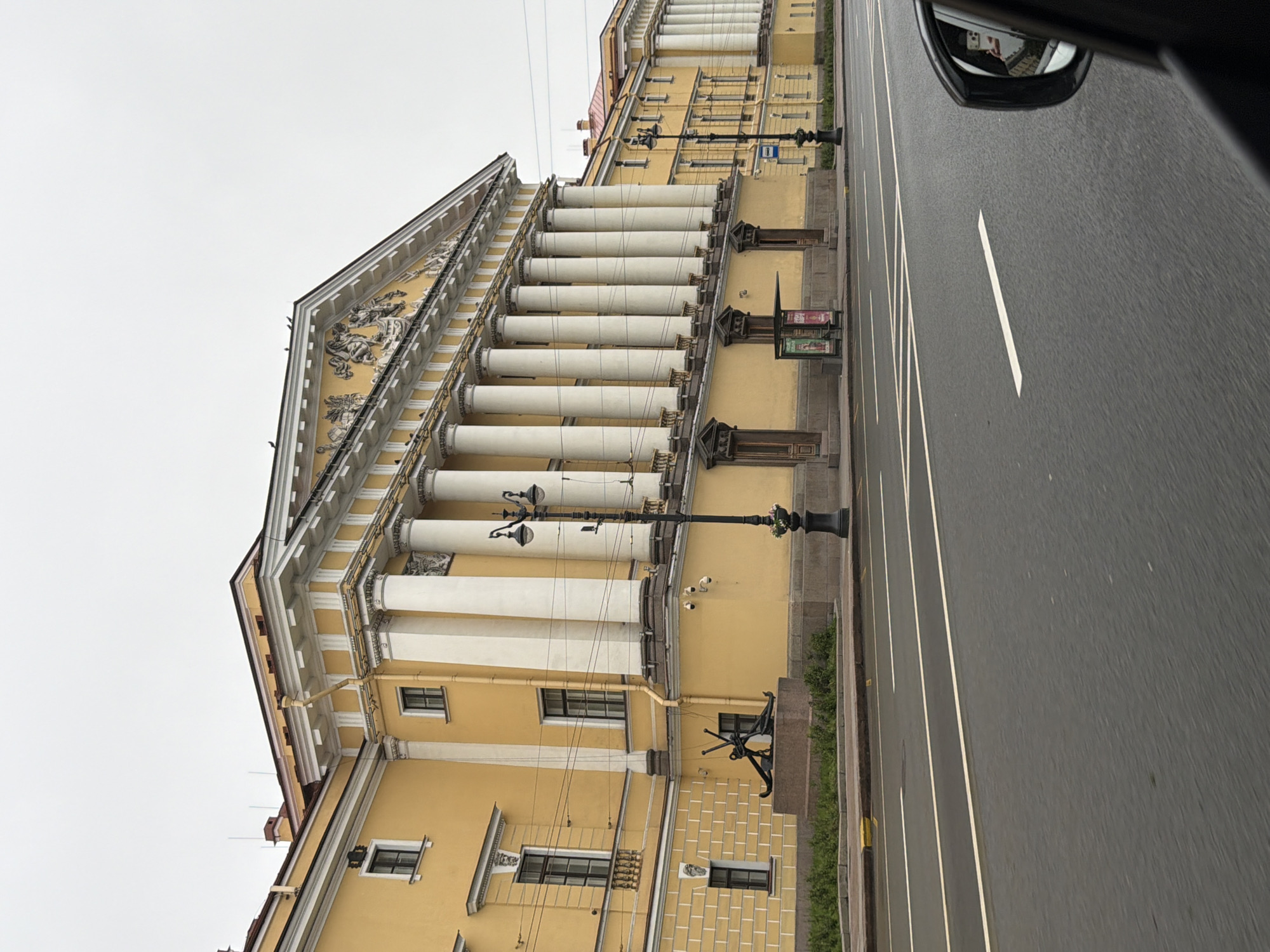 The image shows a side view of a yellow neoclassical building with white columns and an ornate pediment, located on a street in Saint Petersburg, Russia. The building appears to be a significant architectural landmark. In front of the building, there is a sidewalk with greenery, a stone pedestal, and an anchor sculpture. Across the street, a road with traffic lanes is visible, and a black car's side mirror is partially in the frame, reflecting a phone with a selfie. The sky is overcast, suggesting a cloudy day. The overall impression is of a grand, historic urban setting.