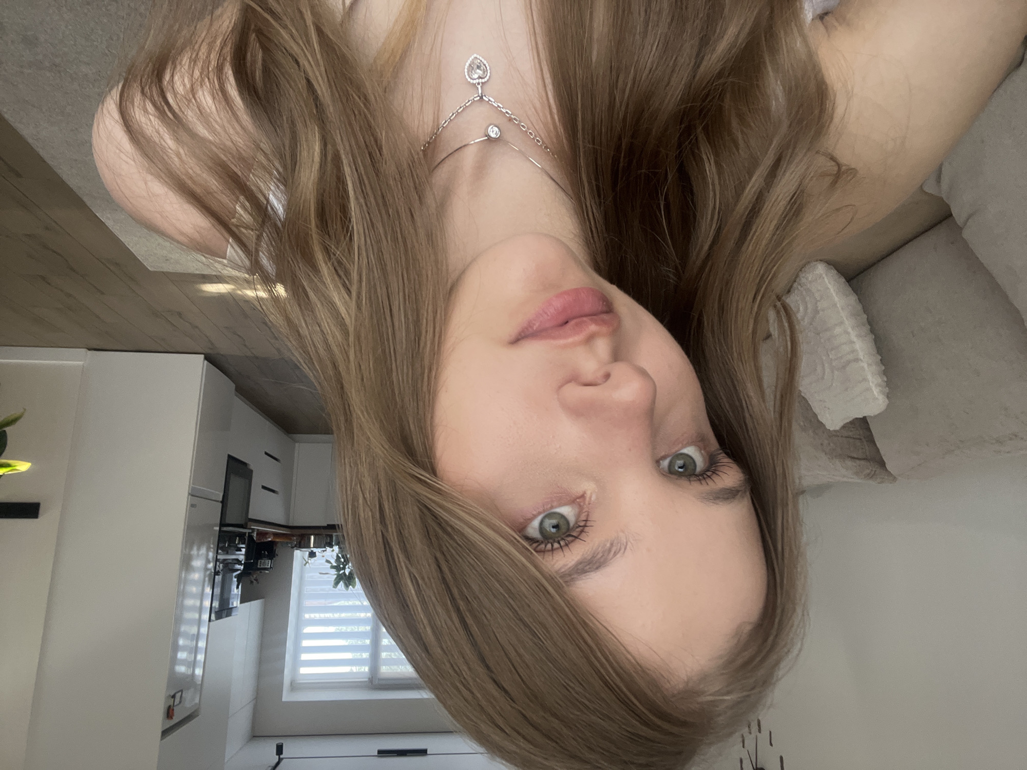 The image depicts a woman with long brown hair, fair skin, and green eyes. She is wearing a delicate silver necklace with a heart-shaped pendant and a secondary design. The woman appears to be lying down, as the background includes a white couch and part of a kitchen with white cabinets, a microwave, and a window with blinds. The image is rotated, giving an upside-down perspective. The location in the city of Votkinsk, Russia, cannot be determined from the given image.