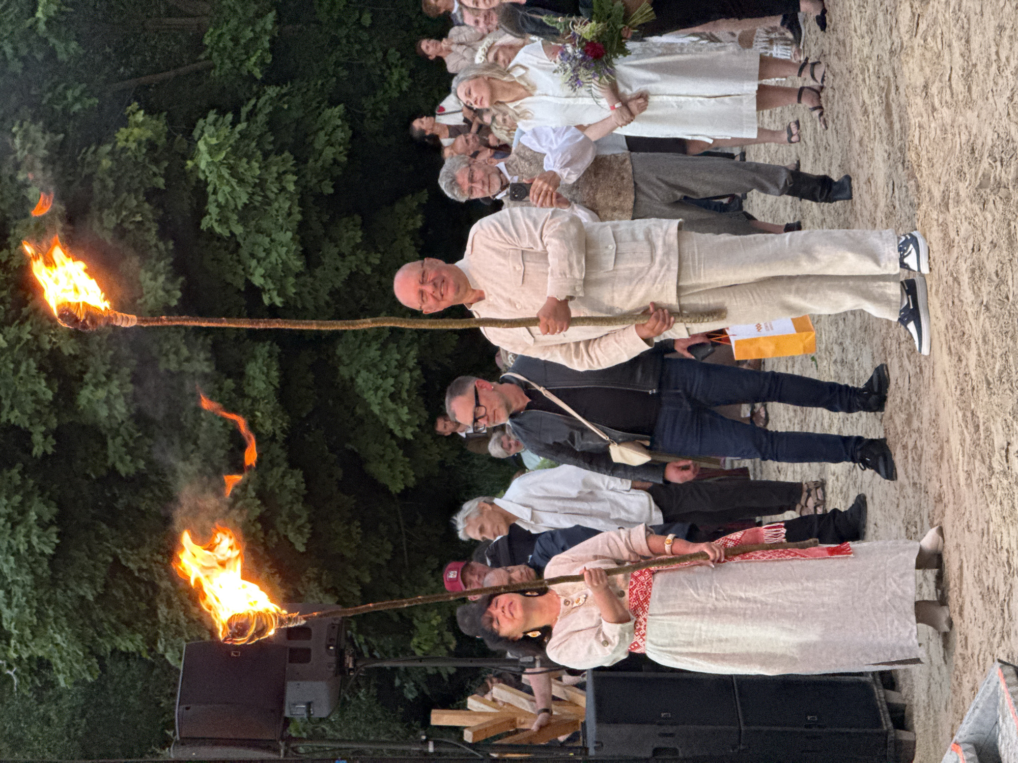The photograph captures a torch-lit outdoor gathering on a sandy surface, likely a beach, with lush green trees forming the backdrop. In the foreground, a woman on the left holds a lit torch, her face tilted upwards. She is adorned in a light-colored, flowing dress with a red and white patterned sash around her waist. To her right, a man in a light-colored suit stands holding another lit torch. Behind them, a crowd of people are visible, some holding phones, suggesting they are documenting the event. A man with glasses in a black leather jacket is also present in the middle ground, to the left of the man with the torch. The atmosphere appears festive and ceremonial. The presence of a sound system speaker on the left edge of the frame suggests a performance or organized event is taking place. The setting, particularly the sandy ground and natural foliage, is consistent with locations in the city of Jūrmala, Latvia, known for its coastal beaches and natural parks.
