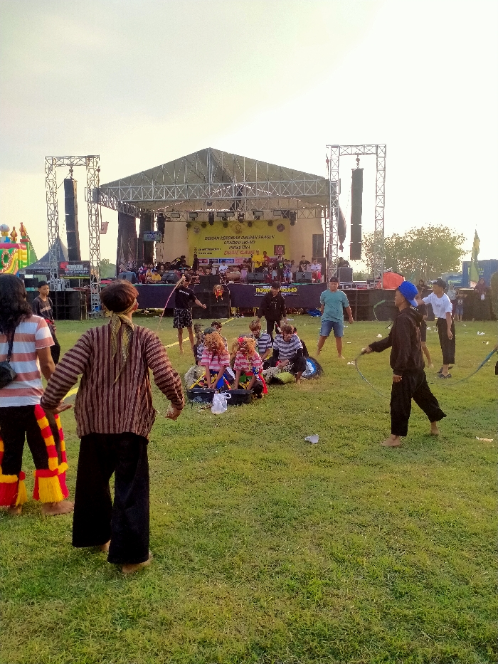 The image depicts an outdoor cultural event in Glonggong, Indonesia, set on a grassy field under a bright sky, likely during late afternoon or early evening. In the background, a large temporary stage constructed from metal trusses with a triangular roof is prominent. Two tall speaker towers with multiple suspended line array speakers flank the stage. A yellow banner with Indonesian text, including "DEWAN KESENIAN DAERAH SRAGEN" (Regional Arts Council of Sragen), "GENDENG RAEAP", "DUSUN GLAWE", and "BUKIT GAWE", is displayed at the rear of the stage, where several individuals are visible, some with musical instruments.
In the foreground and midground, multiple individuals are present on the grass. From left to right: a person with long dark hair wears a striped shirt and traditional red and yellow patterned pants. Beside them, another individual, viewed from the back, wears a vertical striped shirt and dark pants, with a striped bandana on their head. In the midground, a group of approximately five individuals is seated or kneeling on the grass, clustered around several brightly colored traditional horse-like masks (likely part of a Jathilan or Kuda Lumping performance), suggesting preparation or rest. Two distinct masks are visible, one predominantly pink and white, the other blue.
To the right of this group, three standing males are visible: one in a black shirt holding a whip-like object, another in a light blue t-shirt and shorts, and a third in a black hoodie and pants, wearing a blue cap and holding a whip or rope, facing right. The ground is covered in green grass with some scattered white debris. A portion of an inflatable bouncy castle is visible in the far left background.