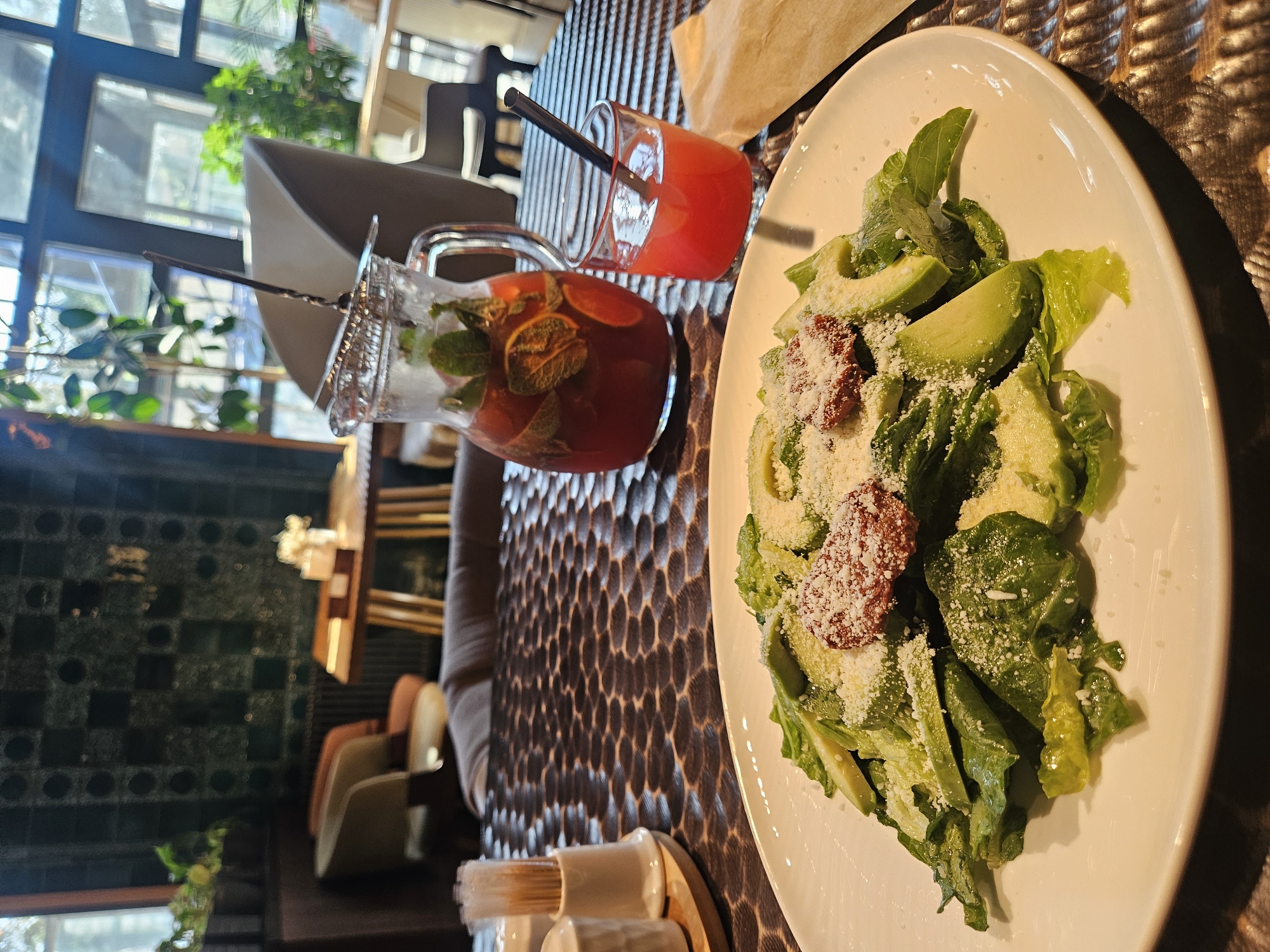The image depicts a table setting with a salad and beverages, likely in a restaurant or café in Malakhovka, Russia. The table has a textured, possibly patterned, surface. A white plate holds a salad consisting of lettuce, avocado slices, and what appears to be crumbled cheese and sun-dried tomatoes. A glass and a pitcher contain red-colored drinks, possibly cocktails or fruit-infused beverages. The pitcher has mint leaves and citrus slices in it. A straw is visible in the glass. Toothpicks in a holder are situated near the table's edge. The background features a window with daylight and glimpses of greenery. Restaurant seating and tiled walls are visible, contributing to the ambiance.