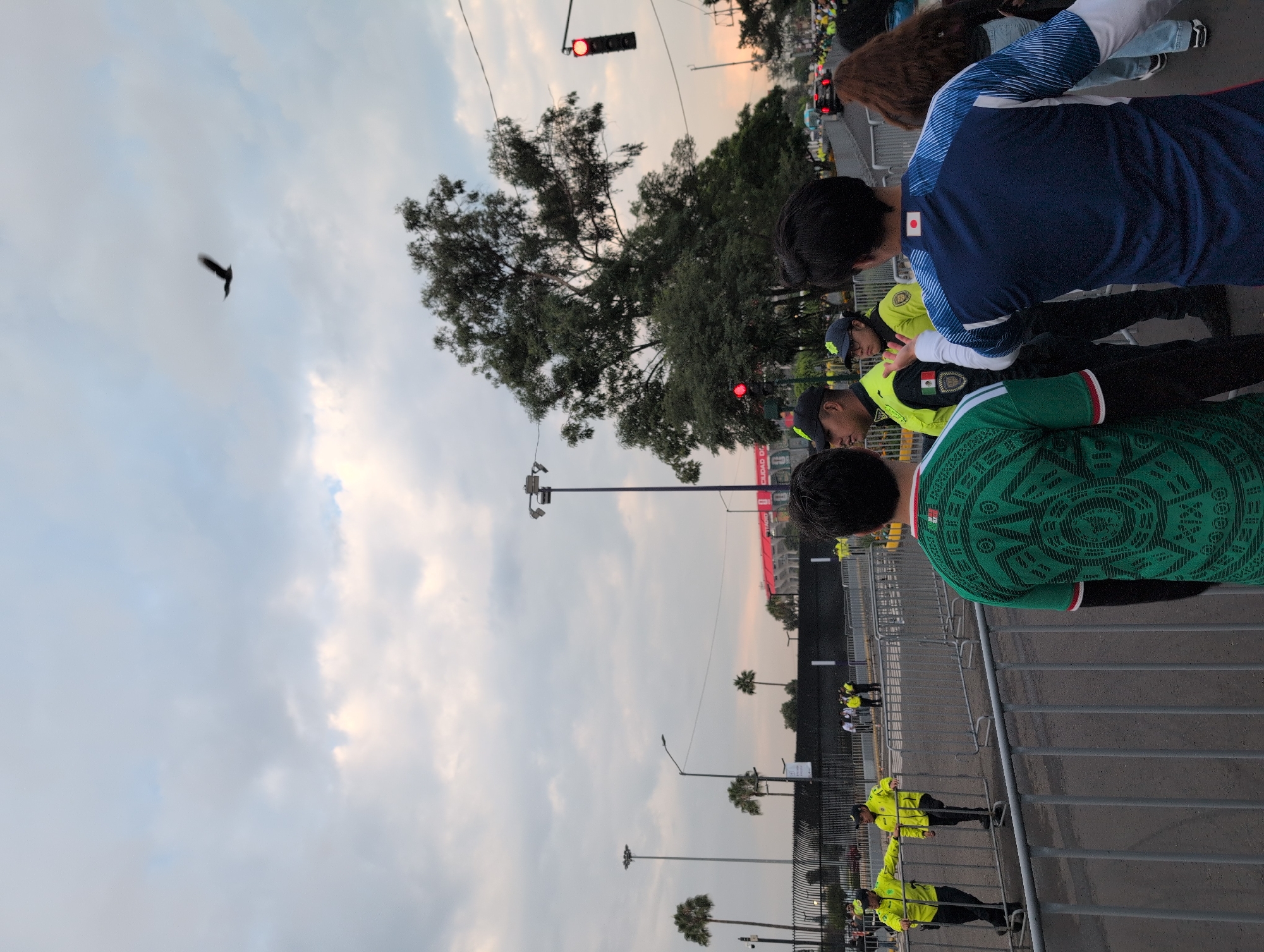 In Mexico City, Mexico, a group of people, some wearing soccer jerseys, stand behind metal barriers. Three individuals in fluorescent yellow vests, possibly security personnel, are visible in the mid-ground. One of them appears to be speaking to another person, who is wearing a dark cap and a matching yellow vest. In the foreground, two young men are seen from behind; one wears a green and white jersey with a geometric pattern, and the other wears a blue and white jersey with a Japanese flag emblem. Further back, a large, leafy tree dominates the right side of the frame, with a traffic light showing red on its right. In the background, a red stadium or building structure is partially visible. The sky is cloudy, and a bird is in flight in the upper right portion of the image.