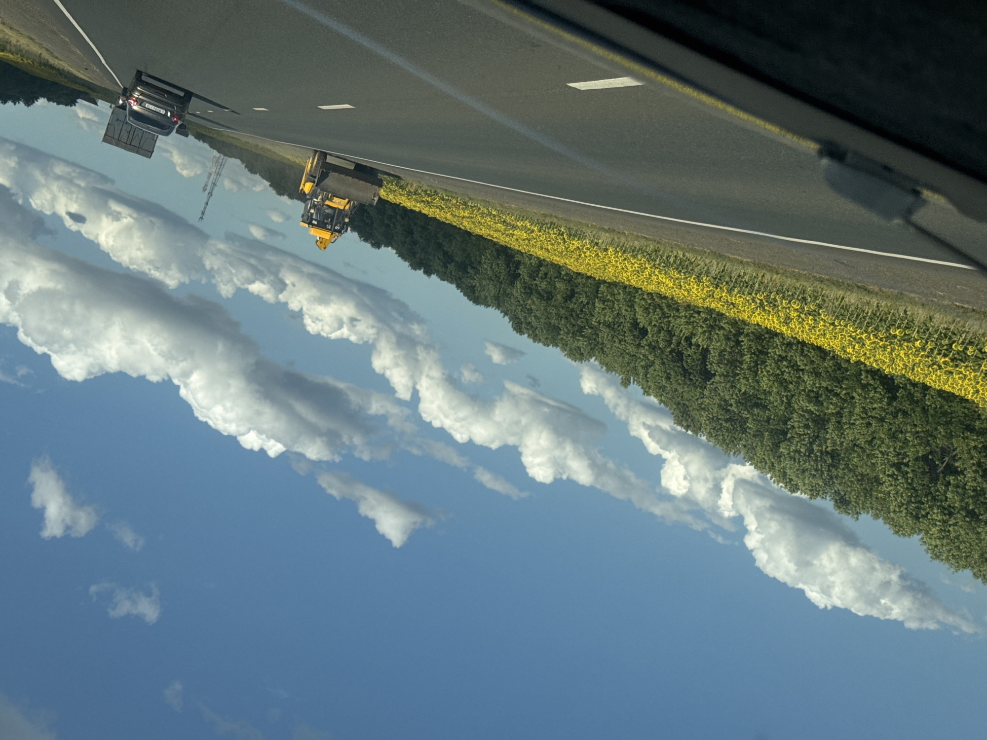 The image depicts a rural highway scene under a partly cloudy blue sky. The viewpoint is from inside a vehicle, looking forward and slightly to the right. On the left side of the highway, a vast field of sunflowers stretches towards a dense line of green trees. The road itself is asphalt with white lane markings.
In the right lane of the highway, moving away from the viewer, is a yellow backhoe loader. Further ahead and to the right, a dark-colored sedan is also traveling in the same direction. Beyond the sedan, a large truck with a trailer is visible. In the distance, a tall telecommunications tower can be seen.
The overall impression is that of a sunny day in a rural area, possibly in the countryside near Perevolotsky, Russia, given the presence of sunflowers, which are commonly cultivated in the region. The scene is devoid of any people visible outside of the vehicles.