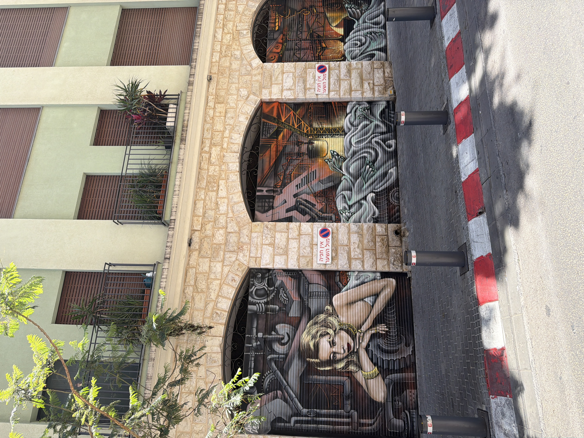 The image shows a three-story building with street art on the ground floor, likely located in Tel Aviv, Israel. The building's first two stories are painted in a light yellow-green with dark brown wooden panels on the windows. The ground floor has three arched openings, each featuring a different mural painted on what appears to be roll-up doors or garage-style doors.
From bottom to top, the murals depict:
1. A woman with blonde hair and golden accessories in a steampunk-style setting with gears and pipes.
2. An industrial scene with stairs, pipes, and a central, open structure, with greyish-white, flowing, organic forms at the edges.
3. A similar scene to the second mural with industrial components and organic forms, but slightly more abstract.
Between each mural, there is a small sign with Hebrew writing and a symbol indicating "no parking." On the building's first and second stories are wrought iron railings.
To the right of the building is a paved street with black bollards and a curb marked with alternating red and white stripes. The street surface transitions to a lighter color in the distance, indicating a possible road or pedestrian area. A tree branch is visible in the foreground on the left side.