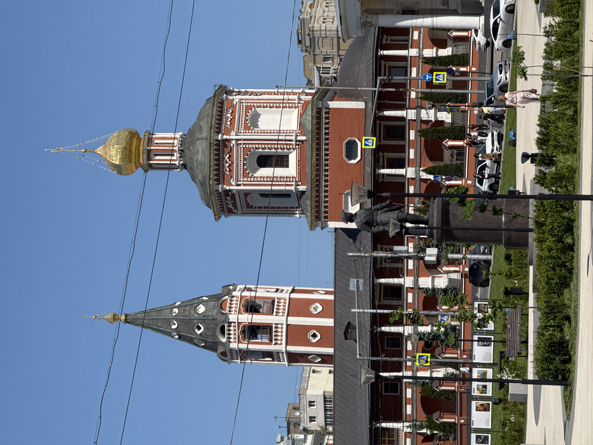 The image shows the red and white facade of a building complex in what appears to be Saratov, Russia. Two distinct towers are visible, one with a golden onion dome and the other with a conical gray roof. The foreground includes a statue, pedestrian crossing signs, benches, and a display of framed images along the walkway. People are present on the sidewalk next to parked cars, and greenery borders the walkway. Overhead wires are visible against the clear blue sky.