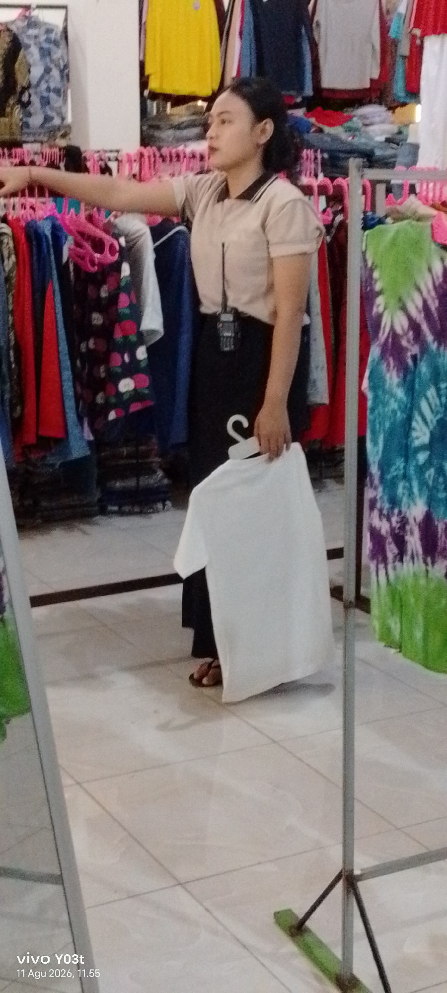 A woman with dark hair pulled back is standing in what appears to be a clothing store. She is wearing a light brown short-sleeved collared shirt and a black skirt. In her right hand, she holds up a white t-shirt on a white hanger. Her left arm is extended, pointing towards racks of colorful clothing in the background. The store features tiled flooring and various clothing items displayed on racks. A device, possibly a radio or pager, is clipped to her shirt. The timestamp on the bottom left indicates the image was taken on August 11, 2026, at 11:55 AM in Atula, Indonesia.