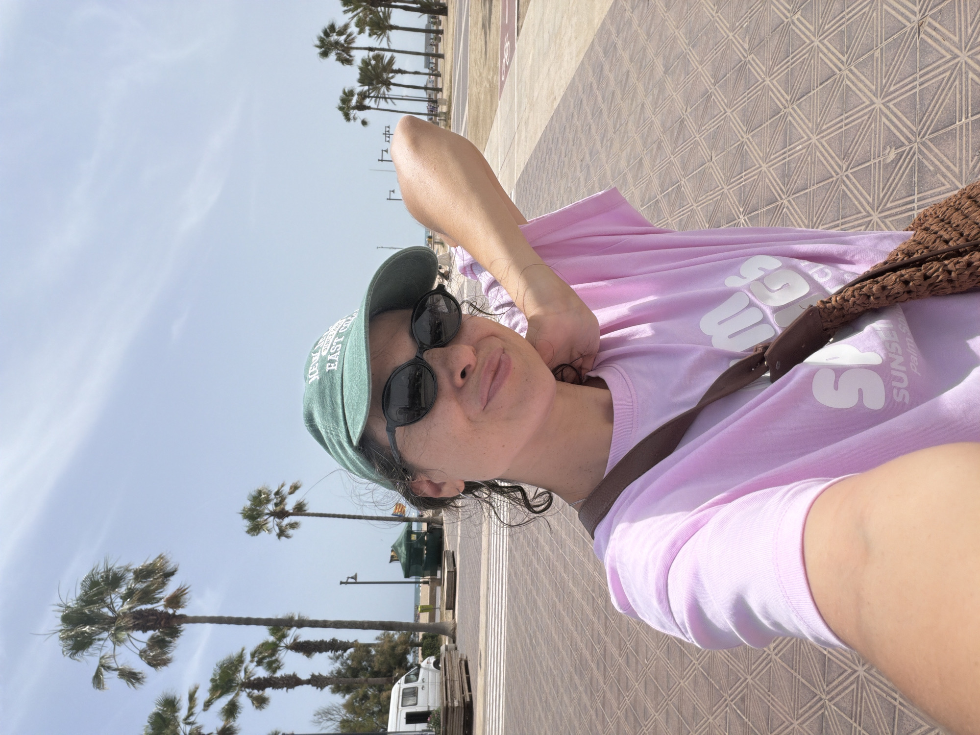The media file is a selfie taken by a person outdoors on a paved walkway lined with palm trees. The person is wearing a pink t-shirt, a green baseball cap, and sunglasses. They are holding their arm up to take the photo, and their hand is touching their neck. A brown woven bag is slung over their shoulder.
The background shows a clear blue sky with some clouds, and several tall palm trees. Further in the distance, there are buildings and a body of water, suggesting a coastal location. The walkway has a distinctive geometric pattern.
The lighting and shadows indicate it is daytime, likely sunny. The overall mood appears casual and relaxed. There is text visible on the person's t-shirt, which reads "SUNSET" and "PALM SPRINGS" in white lettering. There is also text on the person's cap, which appears to be a logo or brand name.
