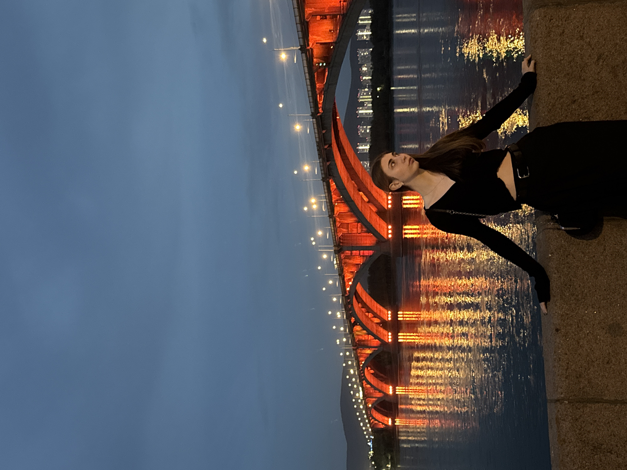 A young woman stands on a stone embankment at dusk, leaning with her arms on the railing. She is wearing a black V-neck top with long sleeves and a black skirt with a belt. A black purse hangs from her left shoulder. Behind her, a large arched bridge, illuminated with vibrant red lights, spans a wide river. The lights of the bridge are reflected in the water, creating a shimmering effect. In the distance, the lights of a city are visible, and the sky is a deep blue, indicating the transition from day to night. The bridge is the Komarovsky Bridge in Krasnoyarsk, Russia, a prominent landmark known for its distinctive illumination. The scene is tranquil, with the woman appearing contemplative as she gazes into the distance.
