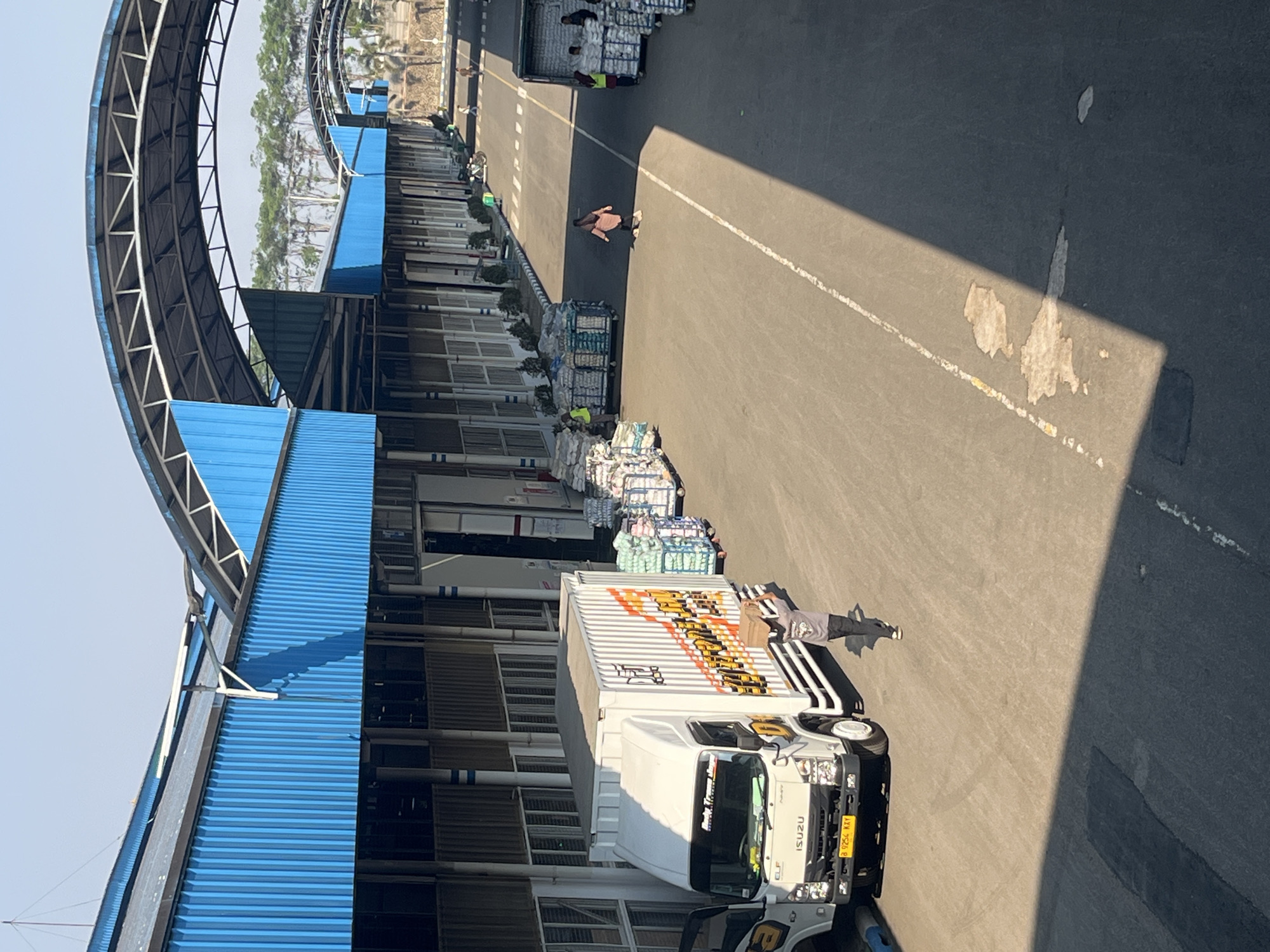 The image depicts an outdoor loading area outside a warehouse with a blue corrugated metal roof structure. Several white trucks, including an Isuzu ELF truck with its door open, are parked on the asphalt. Piles of stacked goods, possibly sacks or bales, are arranged on wheeled carts throughout the scene. People are visible engaged in activities related to loading or unloading. One person, wearing a grey shirt and dark pants, is carrying a cardboard box on their head in the foreground. Another person in a pink headscarf and grey pants is walking away from the trucks further down the road. Several other individuals in vests and casual clothing are also present, attending to the cargo. The environment appears to be a logistical hub or port, with trees visible in the background. The location is identified as Pelang, Indonesia.