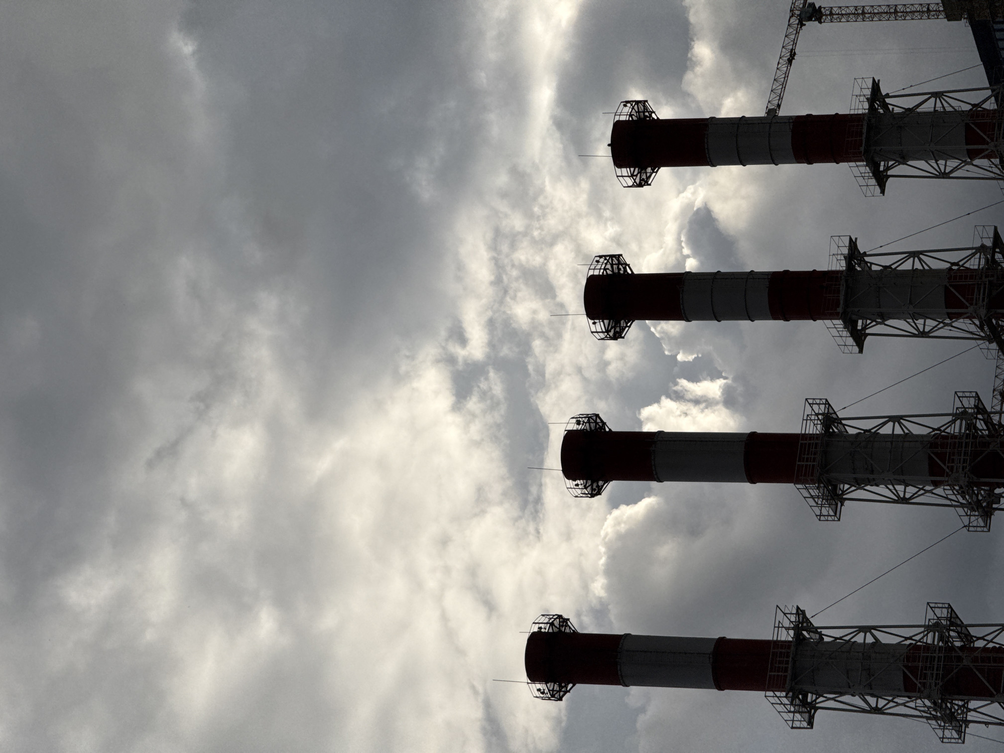The image shows a low-angle shot of four industrial smokestacks against a cloudy sky. The smokestacks are tall and cylindrical, painted with alternating red and white bands. They are supported by intricate metal scaffolding. In the upper right corner of the image, a construction crane is visible, also silhouetted against the sky. The clouds are dense and grey, suggesting overcast weather. The overall impression is one of industrial power and the vastness of the sky. There is no visible text in the image. The lighting suggests it is daytime, but the cloud cover obscures direct sunlight. The scene is likely an industrial area or power plant.