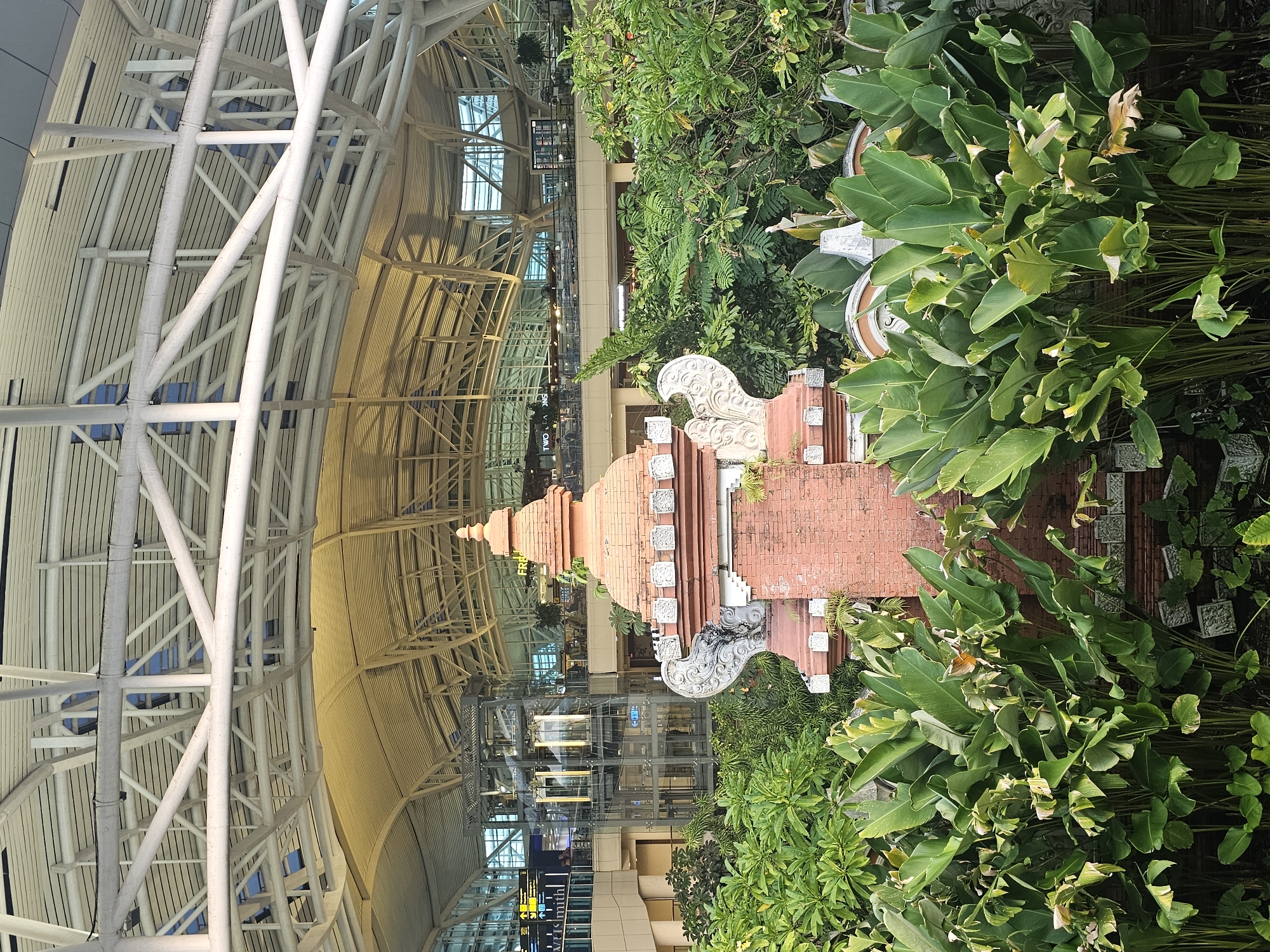 The image depicts an interior scene at Ngurah Rai International Airport (DPS) in Kuta, Bali, Indonesia. A multi-tiered, brick-red Balinese shrine or statue is surrounded by lush green tropical plants. The background reveals the airport terminal interior, including the high-ceilinged architecture with white metal support beams, glass windows, and multiple floors visible through elevators. The environment is bright, well-lit, and populated with passengers and shops in the distance.