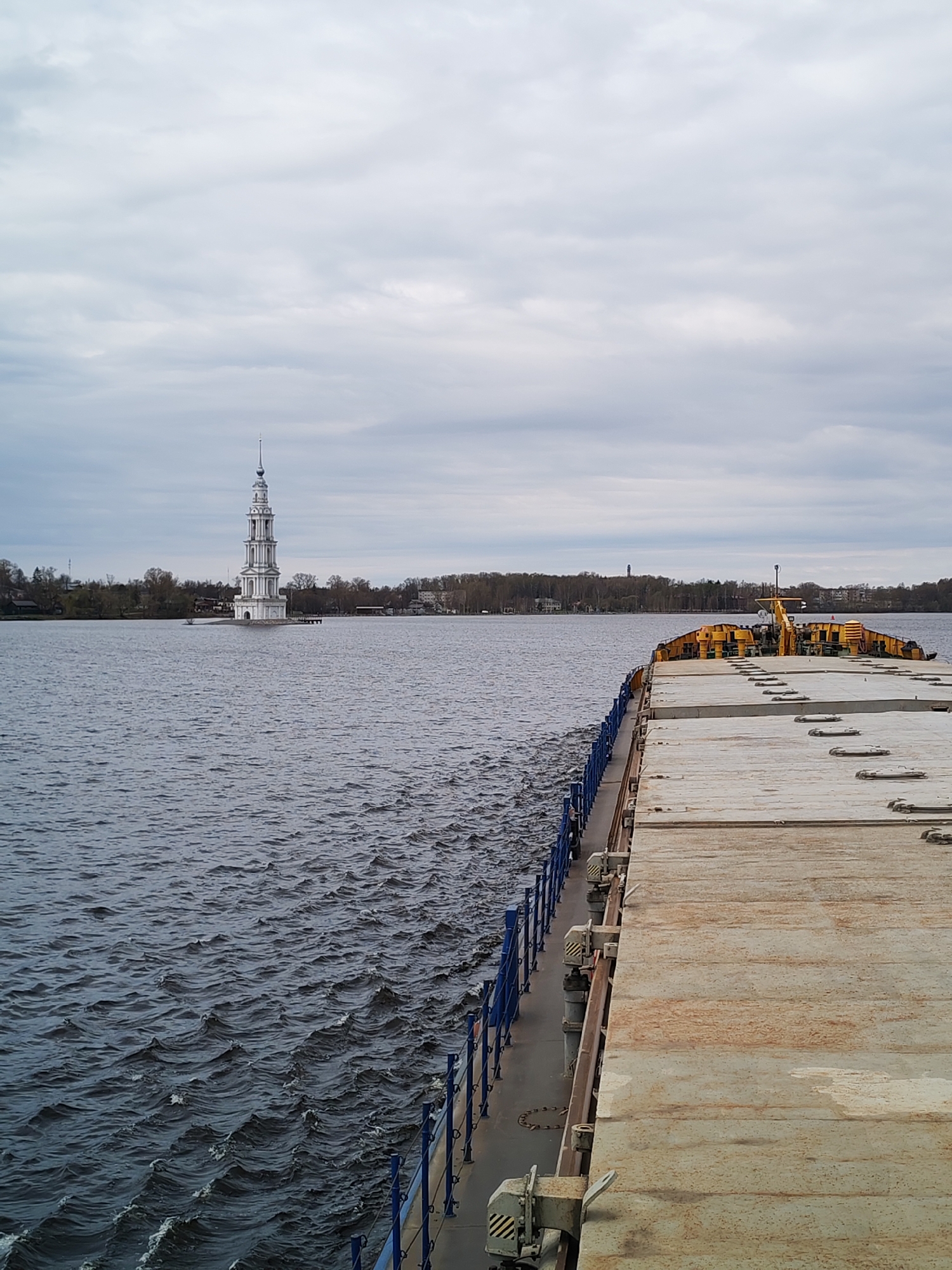 The image depicts a barge on the Volga River in Kalyazin, Russia, with the flooded bell tower of St. Nicholas Cathedral visible in the distance. The bell tower, a prominent landmark, is a white, multi-tiered structure standing in the water. The barge occupies the right side of the frame, showing a flat, concrete-like surface and a blue metal railing along the edge. The water is choppy, with small waves. The sky is overcast with gray clouds. In the background, beyond the bell tower, is the shoreline with trees and buildings. There are no visible people in the image.
