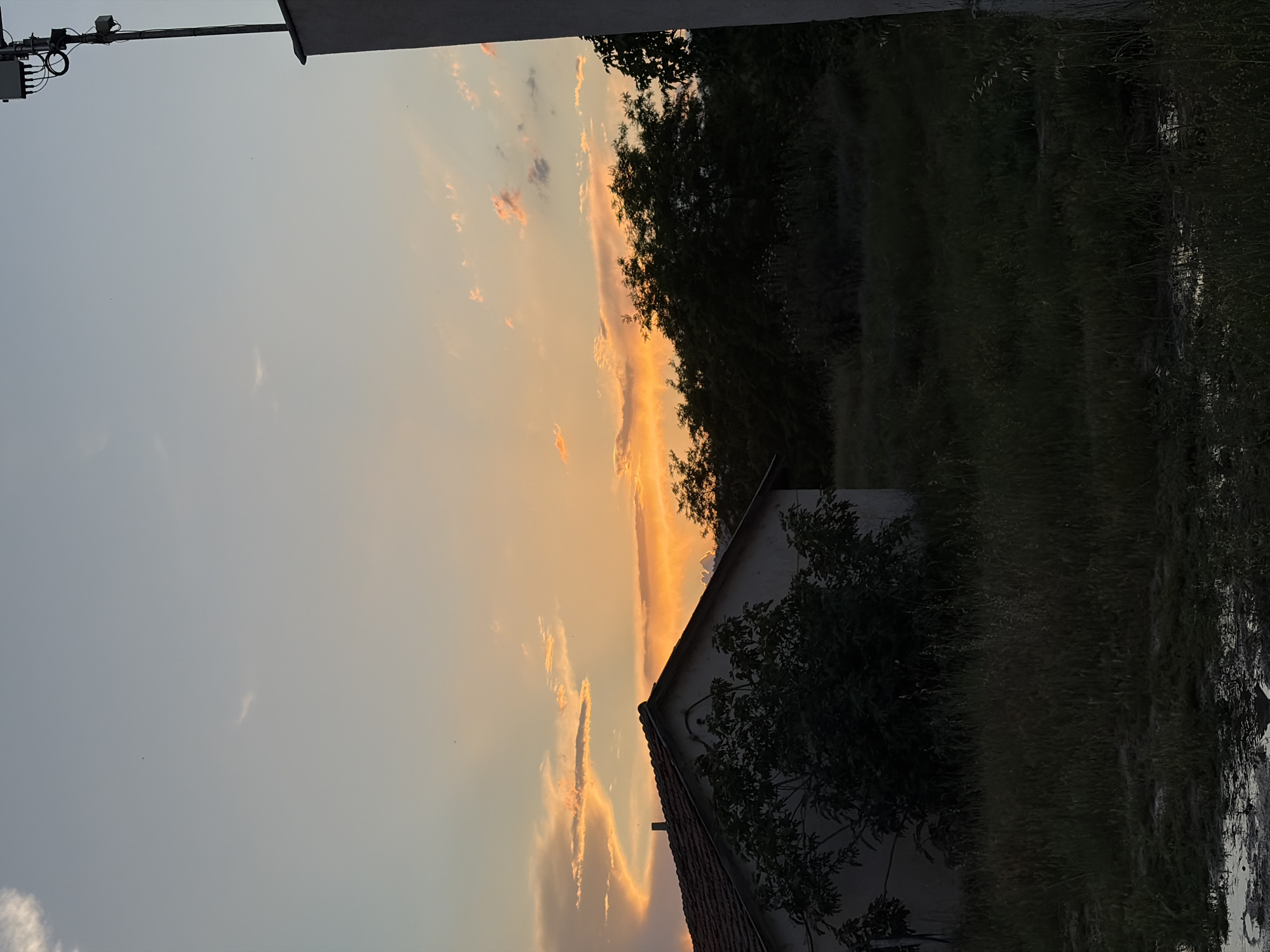 The image depicts an outdoor scene at sunset. The sky is filled with streaks of orange and grey clouds, indicating the time of day. A building with a tiled roof and a tree partially obscures the view of the sunset. Tall grass or foliage is visible to the right of the building, and dark trees or bushes are present in the background. A utility pole with electrical equipment extends from the top left corner of the image. The location is in a city of Unknown, Unknown.