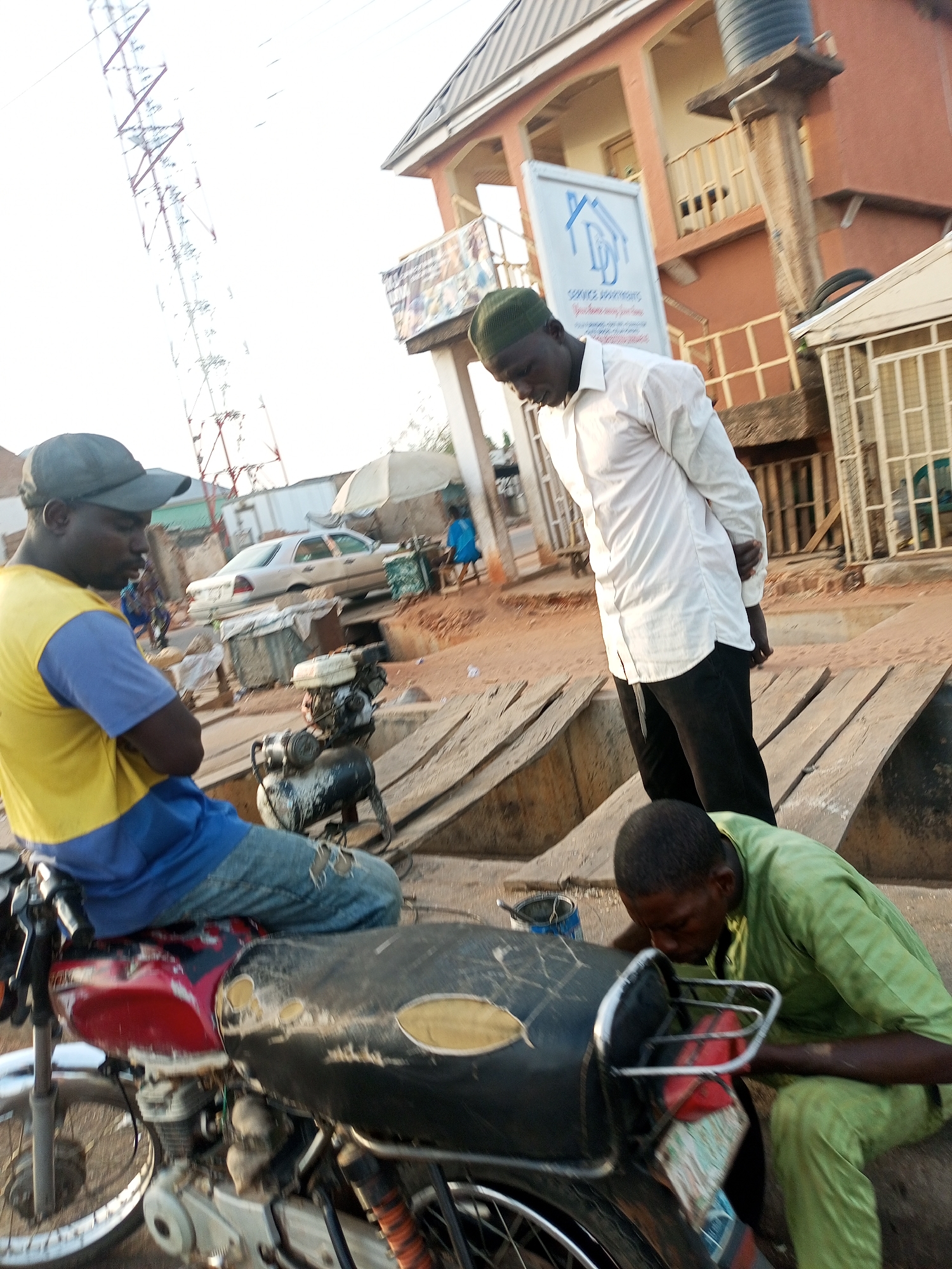 The image captures an outdoor scene in what appears to be Tempure, Nigeria. Three men are present. One man, wearing a blue and yellow shirt and a cap, is sitting on a red and black motorcycle. Another man, dressed in a white shirt and black pants with a green cap, is standing near the motorcycle looking down. The third man, wearing a green shirt and pants, is seen working on the front of the motorcycle. In the background, there is a building with a sign that reads "Service Apartments", and a communication tower is visible in the distance. A car is parked nearby. The ground is dusty, with wooden planks creating a makeshift walkway. Various objects are scattered around, including a small engine and a paint bucket.