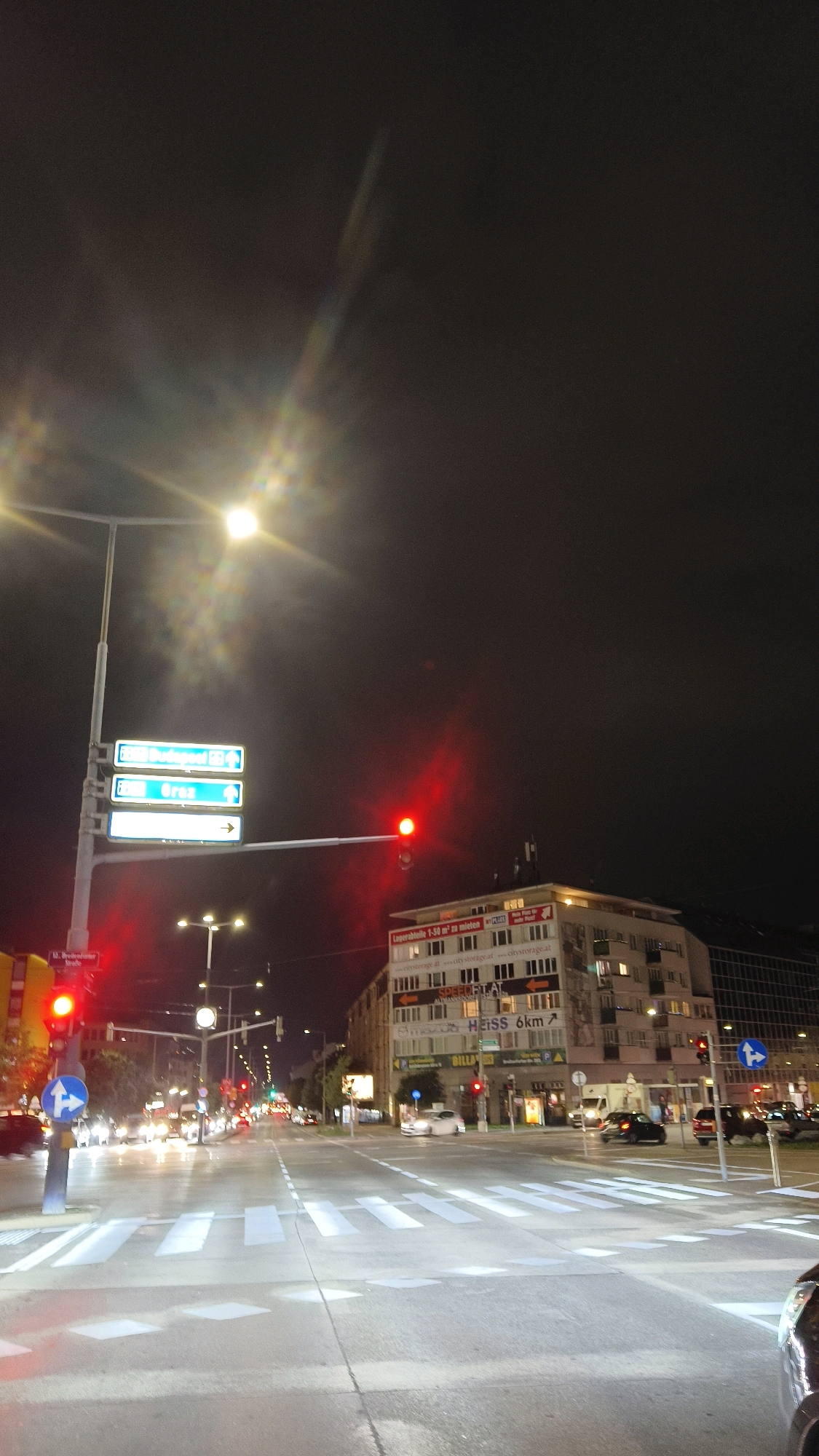 The image captures a nighttime street scene in Vienna, Austria, likely at an intersection. A prominent traffic light shows a red signal, indicating a stop. Several vehicles are visible on the wet road surface, reflecting the streetlights and traffic signals. Buildings line the street, with illuminated windows suggesting occupancy. Signage on one building indicates "HEISS 6km" and references "city storage." Directional signs on a lamppost point towards "Budapest" and "Graz." The sky is dark, with a visible light artifact or lens flare in the upper portion of the frame. Pedestrian crossings are marked on the asphalt.