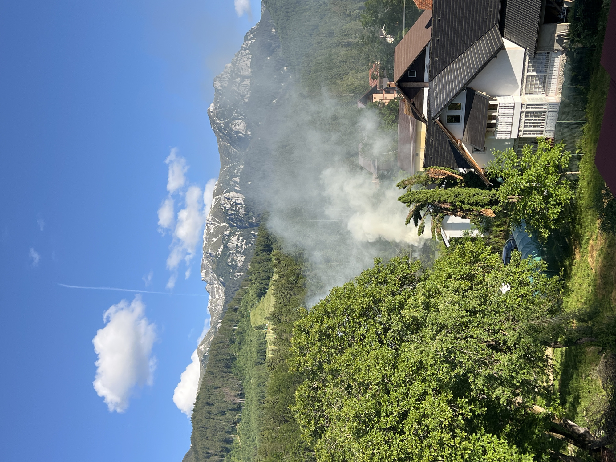 The image shows a residential area with a large mountain range in the background. A significant amount of white smoke is rising from a cluster of trees and homes, obscuring some of the buildings and the lower slopes of the mountain. The sky is a clear blue with a few scattered white clouds and a faint contrail. The sun appears to be high in the sky, suggesting it is daytime. The foreground is dominated by lush green trees. The scene conveys a sense of an event, possibly a fire or controlled burn, unfolding in a picturesque, mountainous location. There is no visible text in the image.