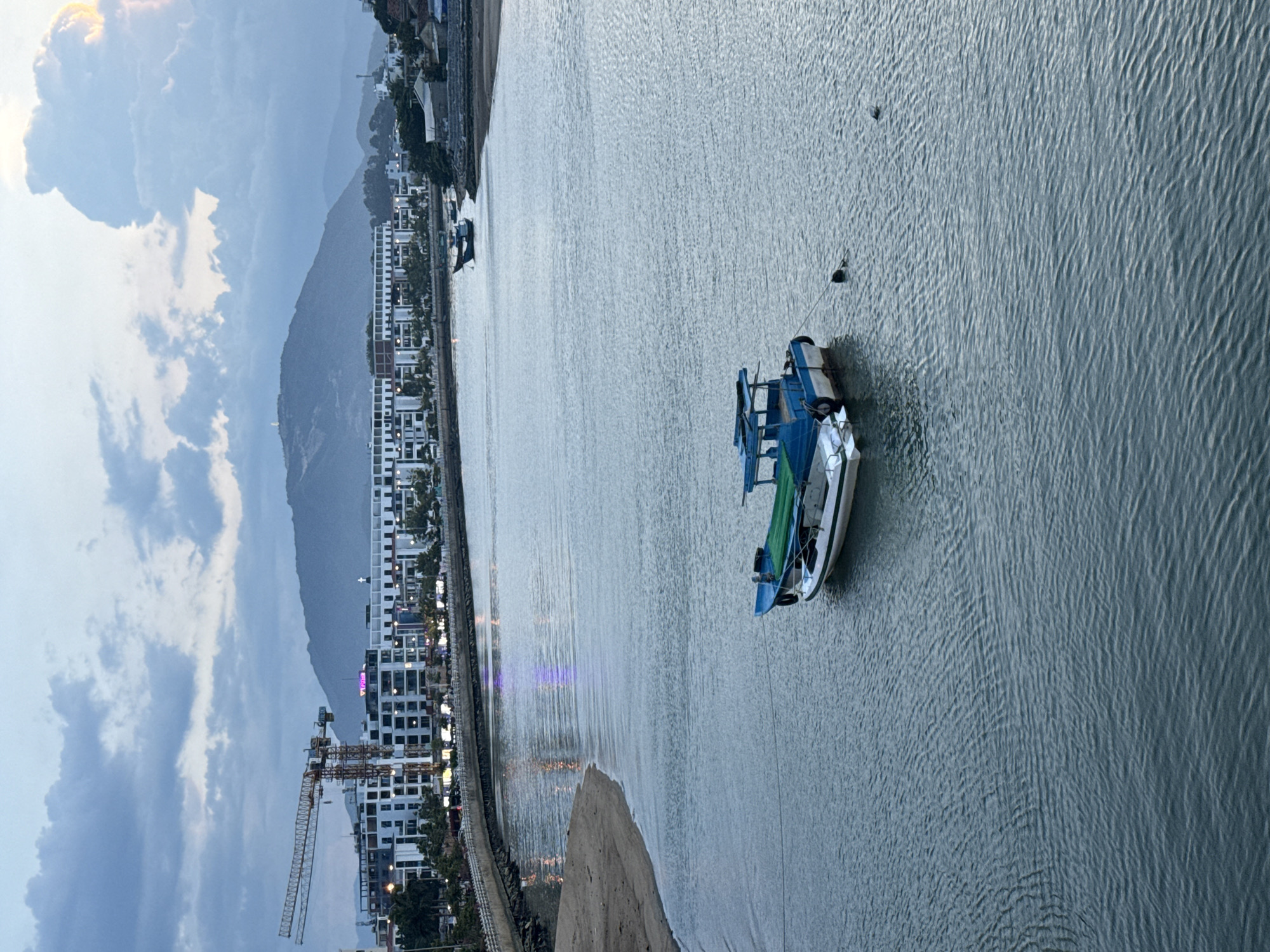 The image depicts a waterfront scene in Nha Trang Ward, Vietnam, during what appears to be twilight or overcast conditions. A calm body of water, likely a river or bay, occupies the majority of the frame. Two boats are visible on the water; one is a small, blue and white vessel with a green tarp on the deck, moored in the foreground. The other boat is further in the distance, also small and dark.
Along the left bank of the water, a sandy beach meets a paved walkway or embankment lined with trees and modern, multi-story buildings. Construction cranes are prominent in the background, indicating ongoing development. The buildings are predominantly white with dark windows and some illuminated signage. Across the water and further inland, a range of hills or mountains forms the backdrop, with some structures and what appears to be a cross visible on the highest peak. The sky is filled with dynamic clouds, some of which are illuminated by soft light, suggesting either sunrise or sunset. A single person or figure, possibly a swimmer or someone in the water, is partially visible near the foreground boat. The overall impression is one of urban coastal development against a natural landscape.
