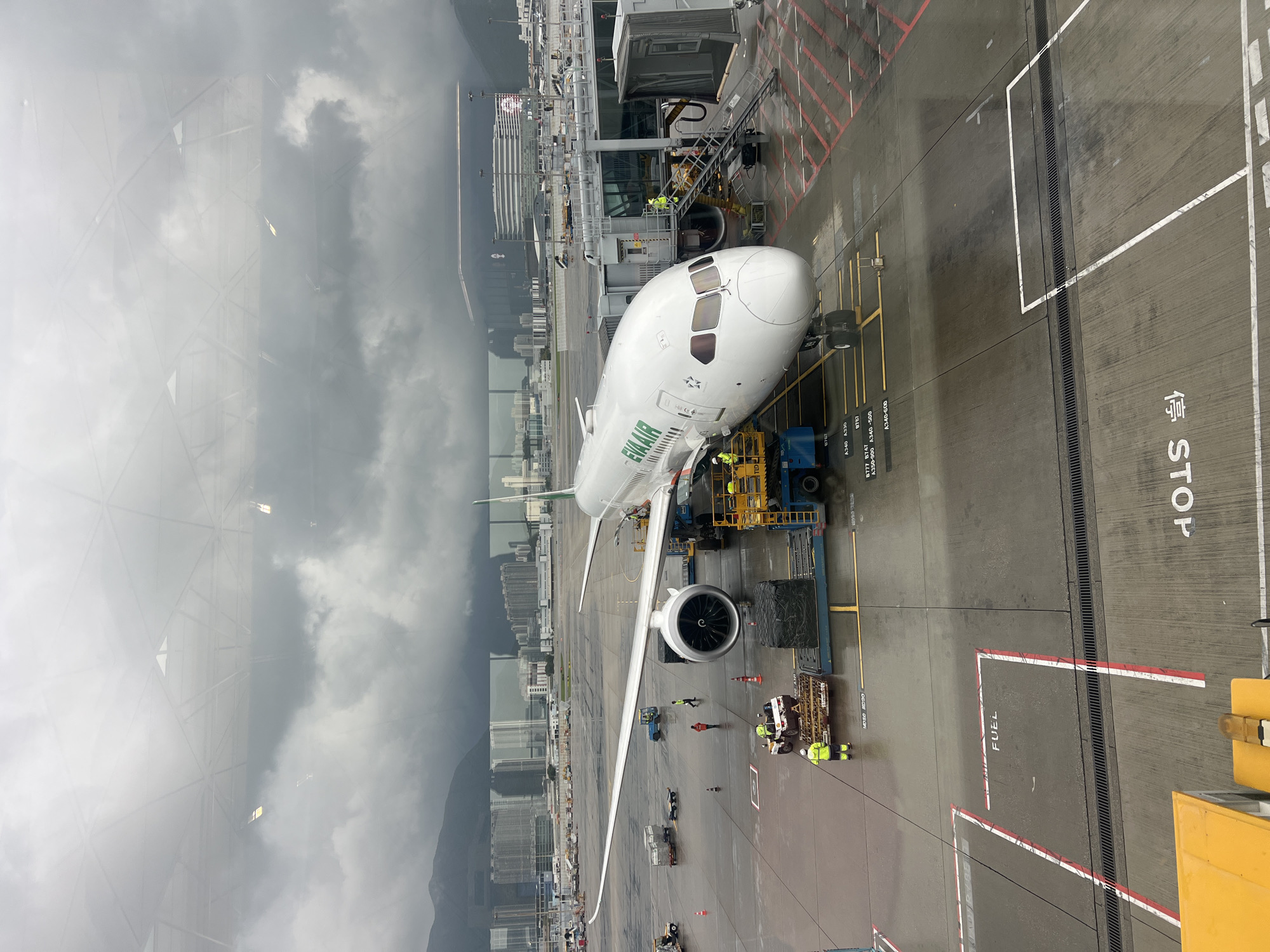 An EVA Air Boeing 787 Dreamliner aircraft, registration B-16701, is parked at a gate at Hong Kong International Airport under a cloudy sky. The plane is white with green and orange accents and the "EVA AIR" logo is visible on the fuselage. The aircraft is connected to a jet bridge, and ground crew members in yellow vests are visible around the plane, some attending to cargo loaders. The airport tarmac is wet, suggesting recent rain. In the background, there is a cityscape with numerous high-rise buildings and mountains, characteristic of Hong Kong. The image is taken from an elevated perspective, likely from within the airport terminal building, looking out through a window that reflects some interior structures.