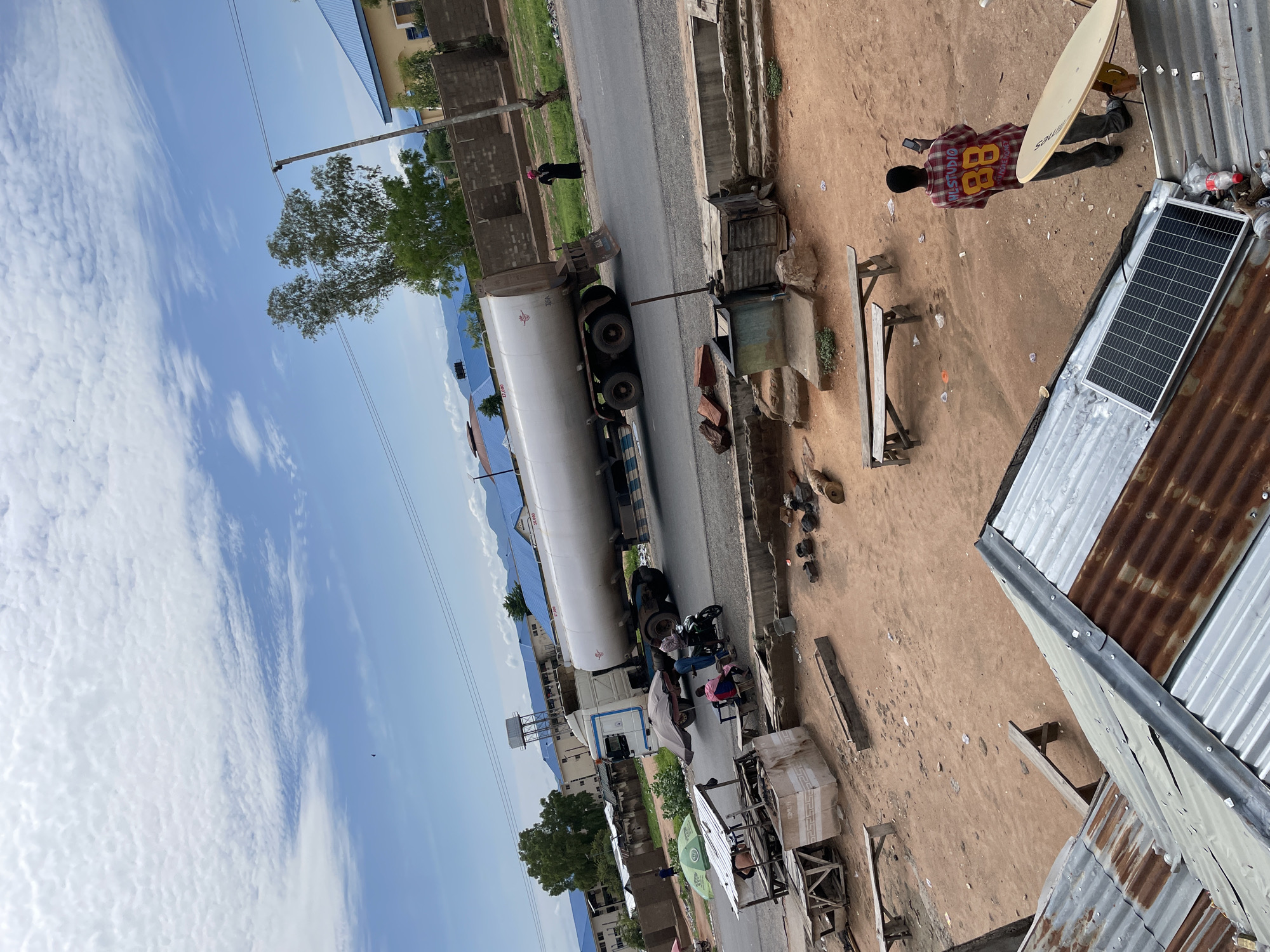 This image captures an outdoor scene in Jalingo, Nigeria, characterized by a mix of commercial activity, residential structures, and an unpaved open area under a partially cloudy blue sky.
The scene is viewed from an elevated perspective, looking down onto a street and an adjacent open dirt lot. In the upper half of the image, a light-colored tanker truck, identifiable by the number "37,000" on its side, is moving from left to right on a paved two-lane road. The truck's cabin is white with blue accents. Several individuals are present near the truck on the roadside. To the left of the truck, under a green market umbrella, are at least two people, one seated at a makeshift wooden stall. Another individual is seated on a wooden bench beside a person on a motorcycle, both seemingly interacting near the side of the road as the truck passes. Further down the road to the right, a lone individual in a dark garment and a pink headscarf stands on the shoulder of the road, observing the street.
The sky is a bright blue with numerous scattered cumulus clouds. In the midground, beyond the road, several buildings with light blue corrugated metal roofs are visible, interspersed with trees. Mountains or hills are faintly visible in the far background.
In the foreground, occupying the lower half of the image, is an unpaved, reddish-brown dirt lot. Various objects are scattered across this area, including several wooden benches, an empty wooden trough, and remnants of what appear to be small fires or discarded items. Partially visible at the bottom of the frame are corrugated metal roofs, some rusted, indicating nearby structures. On one of these roofs, a small solar panel is mounted, and a satellite dish is positioned near the right edge. An individual, seen from behind, is standing in the dirt lot near the satellite dish, holding a mobile phone. This person is wearing a plaid shirt with the number "88" and "W.L. STUDIO" printed on the back.