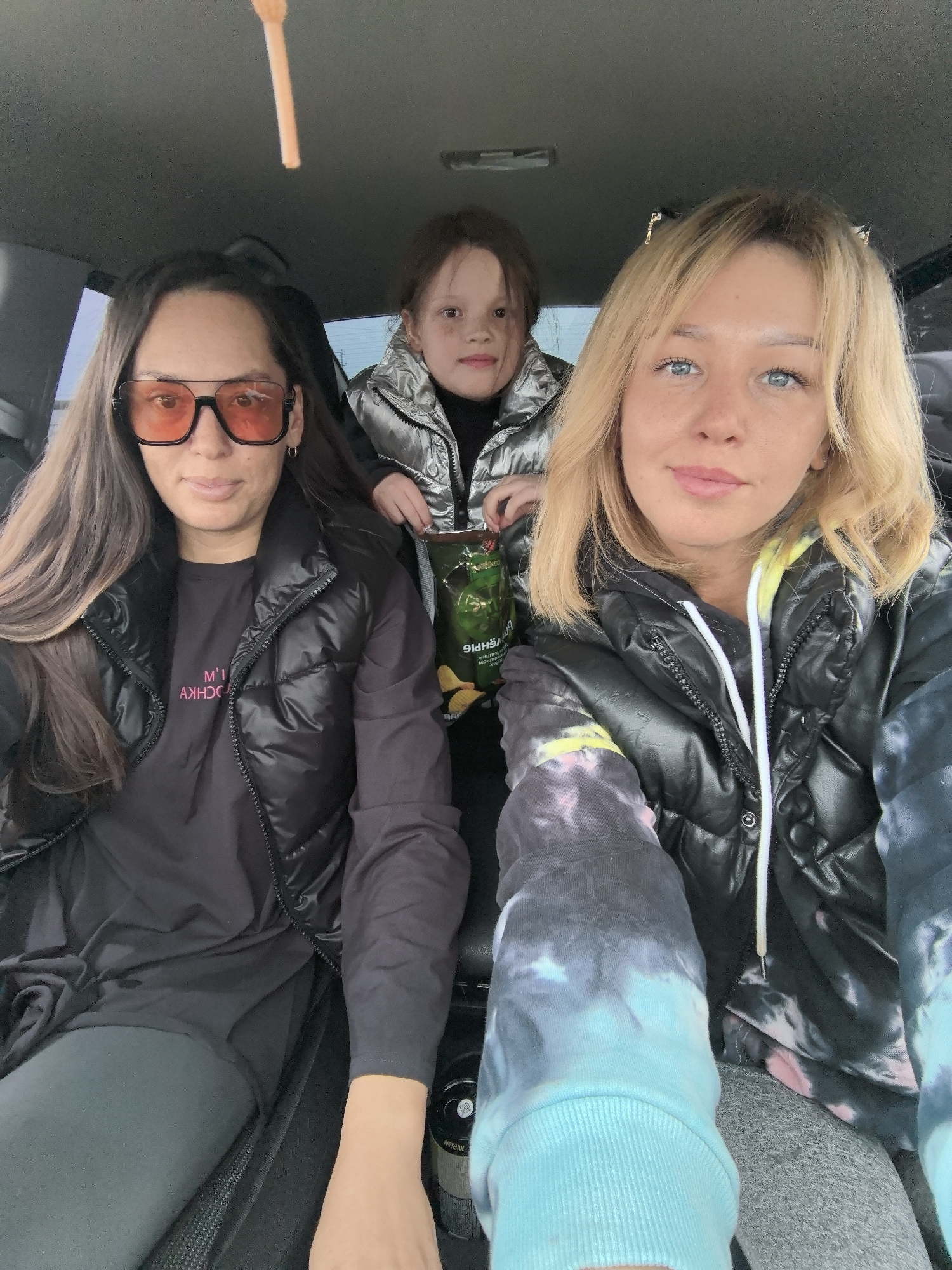 Three people are inside a vehicle. On the left is a woman with long brown hair and dark-tinted glasses. She is wearing a black vest and a dark t-shirt. In the middle is a child in a silver puffer jacket, holding a bag of chips. On the right is a woman with blonde hair and blue eyes, wearing a black vest over a tie-dye hoodie. They appear to be taking a selfie from inside the vehicle. The location is Zav'yalovo, Russia.