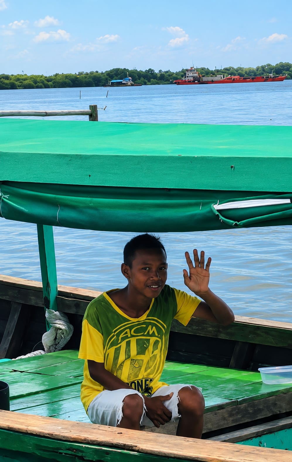 A young boy with short, dark hair and a slight smile is sitting on the green wooden floor of a boat, waving with his right hand. He is wearing a yellow t-shirt with green sleeves and a green collar, featuring a graphic design that includes the letters "ACM" and the word "ONE." His shorts are white and appear torn. The boat has wooden sides and a green canvas covering over the top. In the background, a blue body of water stretches towards a distant treeline. Several boats are visible on the water, including a long orange and white barge and a smaller blue and white vessel. The sky is blue with scattered white clouds. The scene appears to be a waterfront or river area, likely within or near the city of Medan, Indonesia, given the tropical vegetation and the types of vessels present.