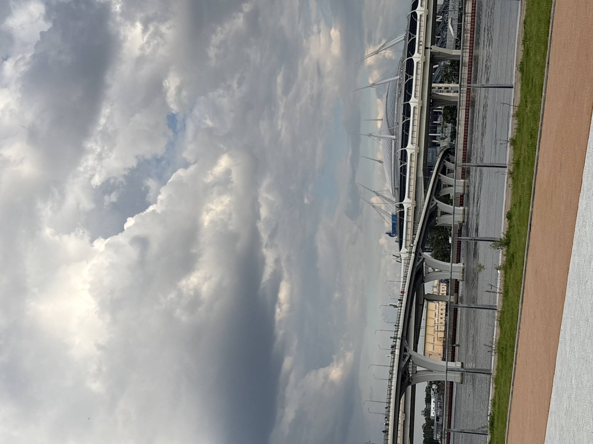 The image captures a panoramic view of a modern urban landscape in Saint Petersburg, Russia, featuring prominent architectural structures under an overcast sky.
The central subject is a large, multi-lane elevated highway or bridge system, likely part of the Western High-Speed Diameter (WHSD), which spans across a body of water. The bridge features numerous concrete pillars and supports, with streetlights lining its edges. To the right, dominating the middle ground, is the distinctive white roof of a large stadium, identifiable as the Gazprom Arena (Krestovsky Stadium). Its futuristic design includes intricate cable-stayed elements and a retractable roof structure.
In the foreground, a paved path, possibly for pedestrians or cyclists, runs parallel to a strip of green grass. The path has a reddish-brown section next to a lighter gray or white section. The body of water beneath the bridge appears calm, reflecting the muted light from the sky. Across the water, distant buildings or industrial structures are faintly visible.
The sky is heavily overcast with dense, grey clouds that fill most of the upper frame. Patches of brighter clouds suggest sunlight attempting to break through, while other areas are darker, indicating thick cloud cover. A small glimpse of blue sky can be seen through a gap in the clouds in one area. The overall weather appears cloudy, but there are no signs of rain.
The time of day is daytime, but the heavy cloud cover gives the scene a muted, somewhat subdued light, making it difficult to determine the exact hour. No clear activity or distinct human figures are prominently visible, and no legible text can be identified on any of the structures. The scene conveys a sense of modern infrastructure set against a dynamic, cloudy sky.