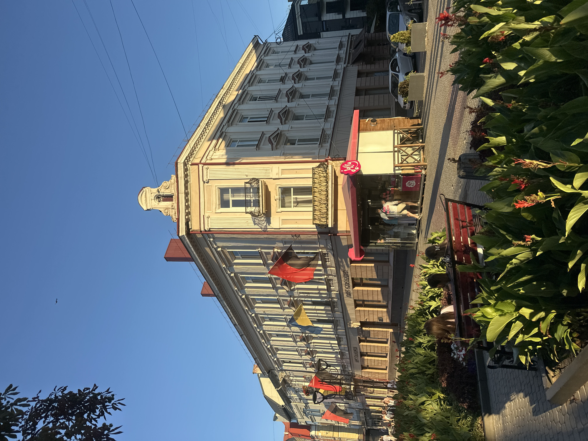 The image depicts a street scene in Ternopil, Ukraine, on a sunny day. A multi-story building with cream-colored facade and red accents dominates the background. The building features classical architectural elements, including decorative moldings, balconies with ornate railings, and numerous windows. Several flags, including the Ukrainian flag, are displayed along the facade. In the foreground, a row of lush green plants with red flowers lines a cobblestone walkway. A red bench is placed among the plants, with several people seated on it. A small kiosk with a pink awning and a pink circular logo is situated on the sidewalk. Several cars are parked on the street, and a few individuals are walking along the sidewalk. The sky is clear blue with a few wispy clouds. The overall atmosphere suggests a pleasant day in an urban environment.