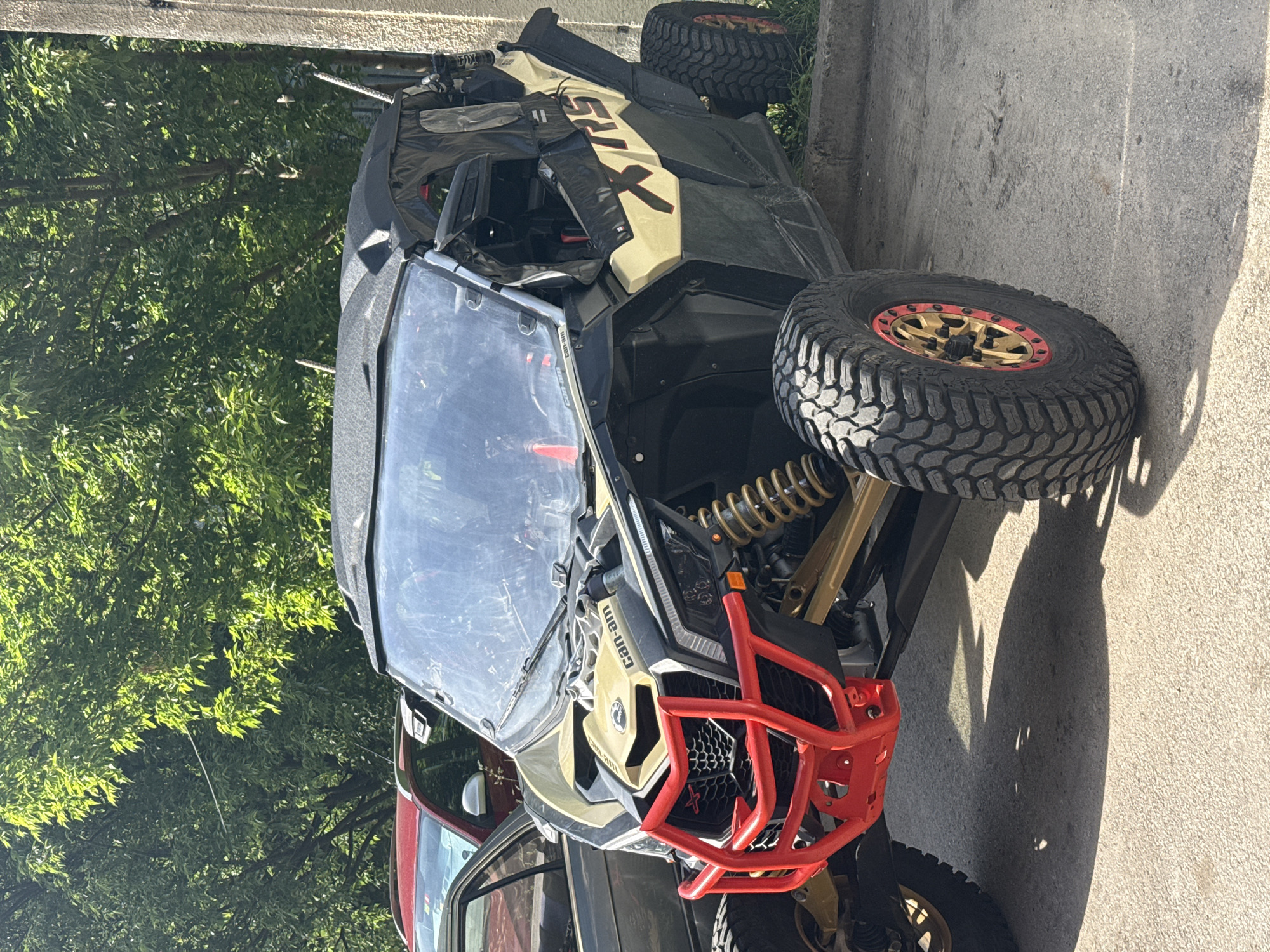 The image shows a Can-Am Maverick X3 side-by-side vehicle parked on a paved surface, possibly a driveway or street. The vehicle is primarily black and tan, with a red front bumper guard. It has large, knobby tires with gold and red rims. The windshield is visible and appears slightly dirty. The background includes green foliage, possibly trees or shrubs. The location cannot be verified from the image.