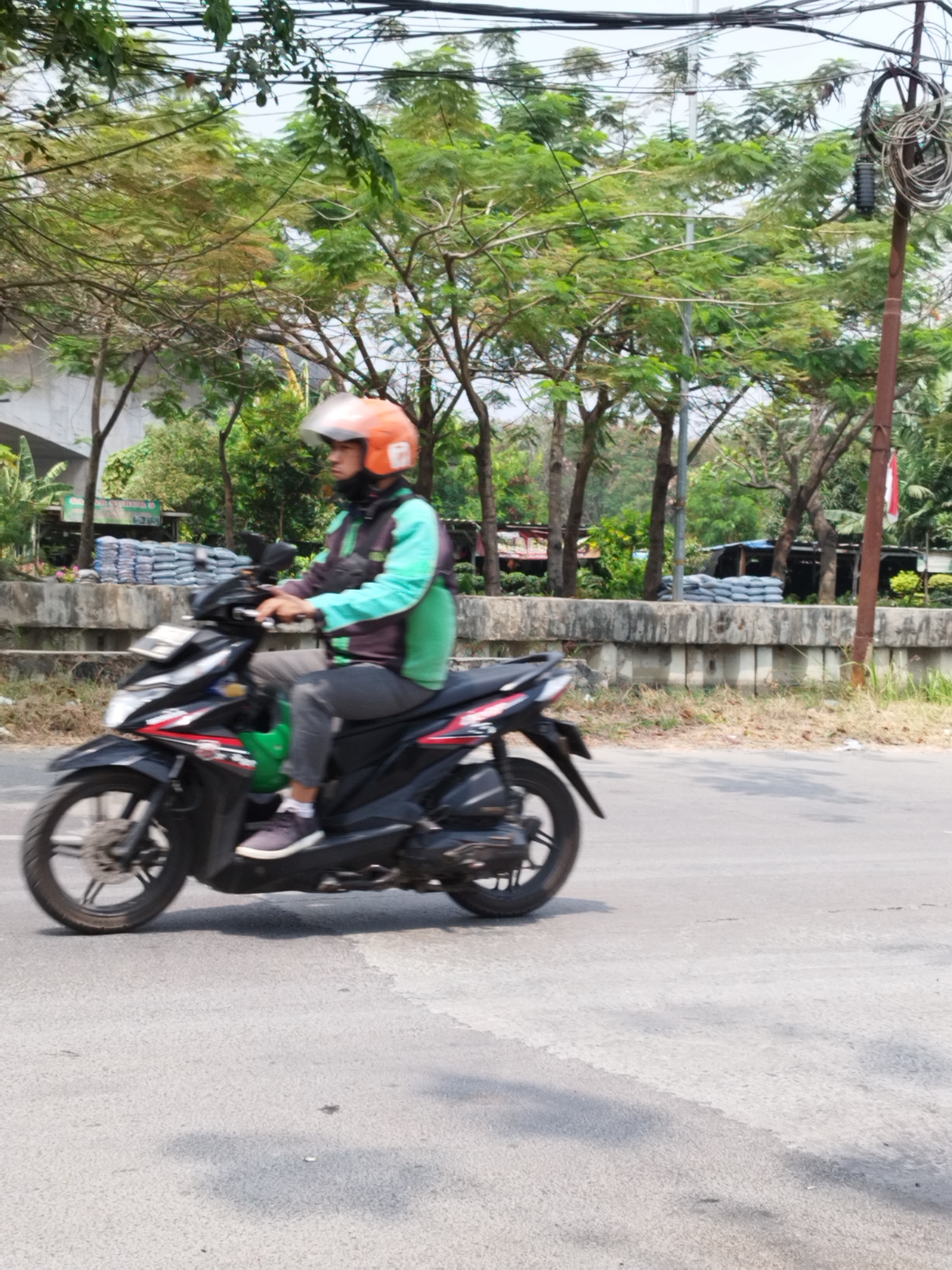 A person wearing an orange helmet and a teal and purple jacket is riding a black motorcycle on a paved road. The motorcycle has red and white accents on its side. The rider's left leg is extended forward, and their right leg is on the footrest. A green bag is hanging from the front of the motorcycle. Behind the motorcycle, there is a concrete barrier with stacks of grey bags piled behind it. Trees with green foliage line the background, and overhead, electrical wires are visible. The scene appears to be outdoors, possibly on a street in Bekasi, Indonesia, given the context.