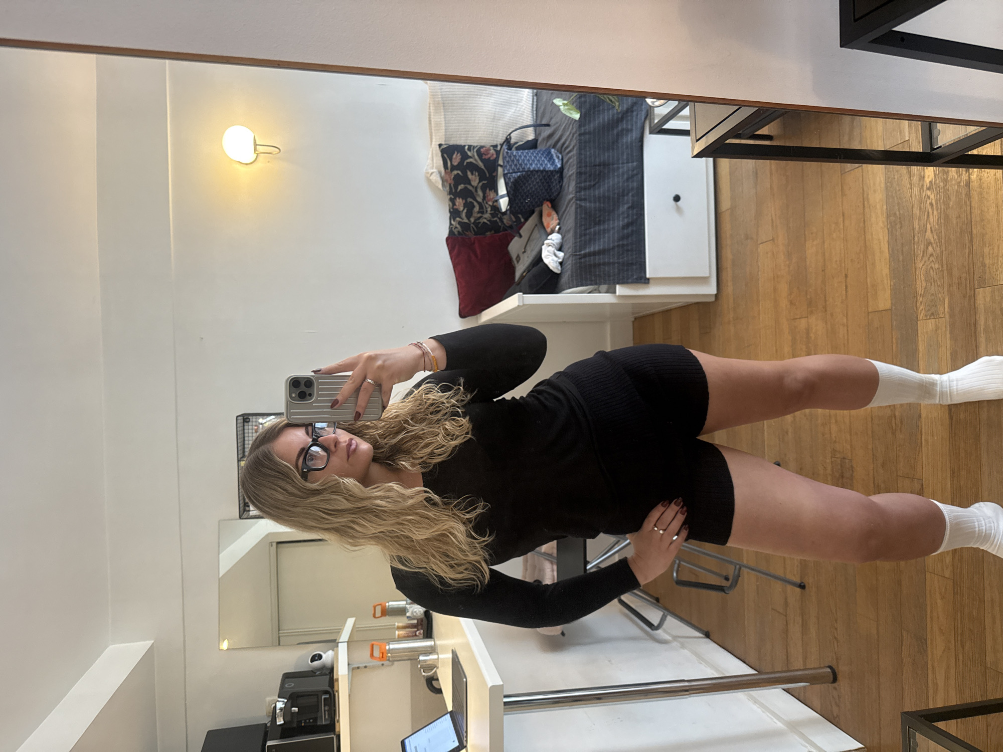 A young woman in a black, long-sleeved dress and white socks is taking a selfie in a mirror. The background shows a room with wooden floors. Reflected in the mirror is a bed with pillows and a blue bag, along with a white desk or shelf unit where a laptop is visible. A small lamp is mounted on the wall. The scene could be an apartment interior. The city of Paris, France cannot be confirmed from the visual data provided.