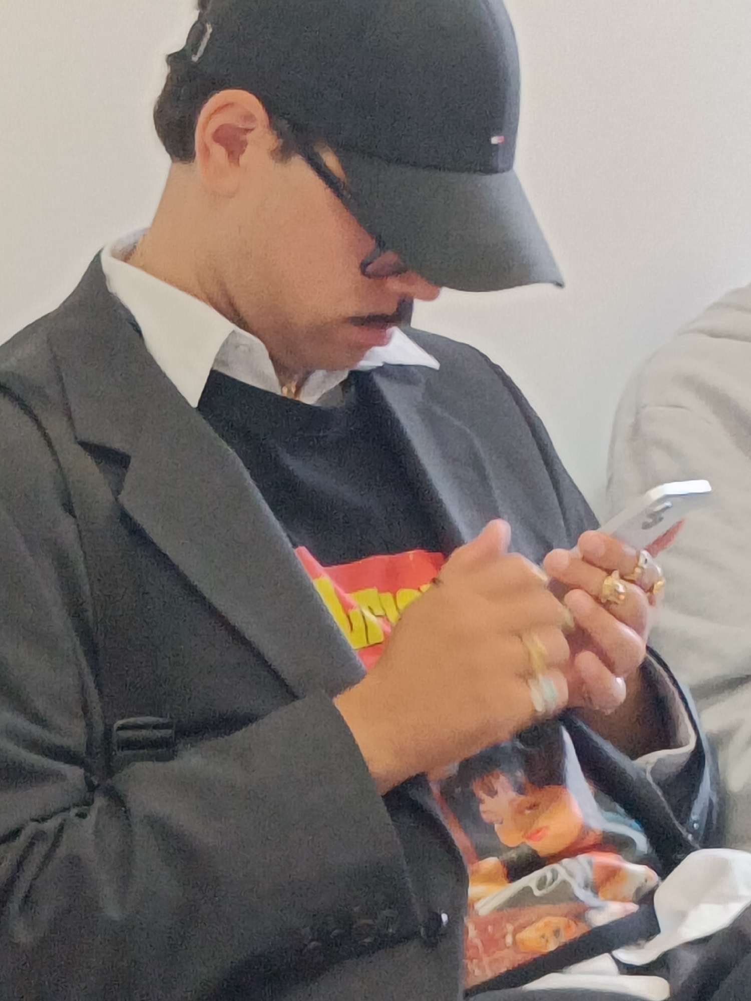 The image shows a close-up, side-profile of a person wearing a black baseball cap, a white collared shirt, and a dark blazer over a graphic t-shirt. The person is looking down at a silver smartphone held in their hands. Several rings are visible on their fingers. The background is a plain, light color. The scene appears to be indoors, possibly in a public setting given the presence of another person in the background.