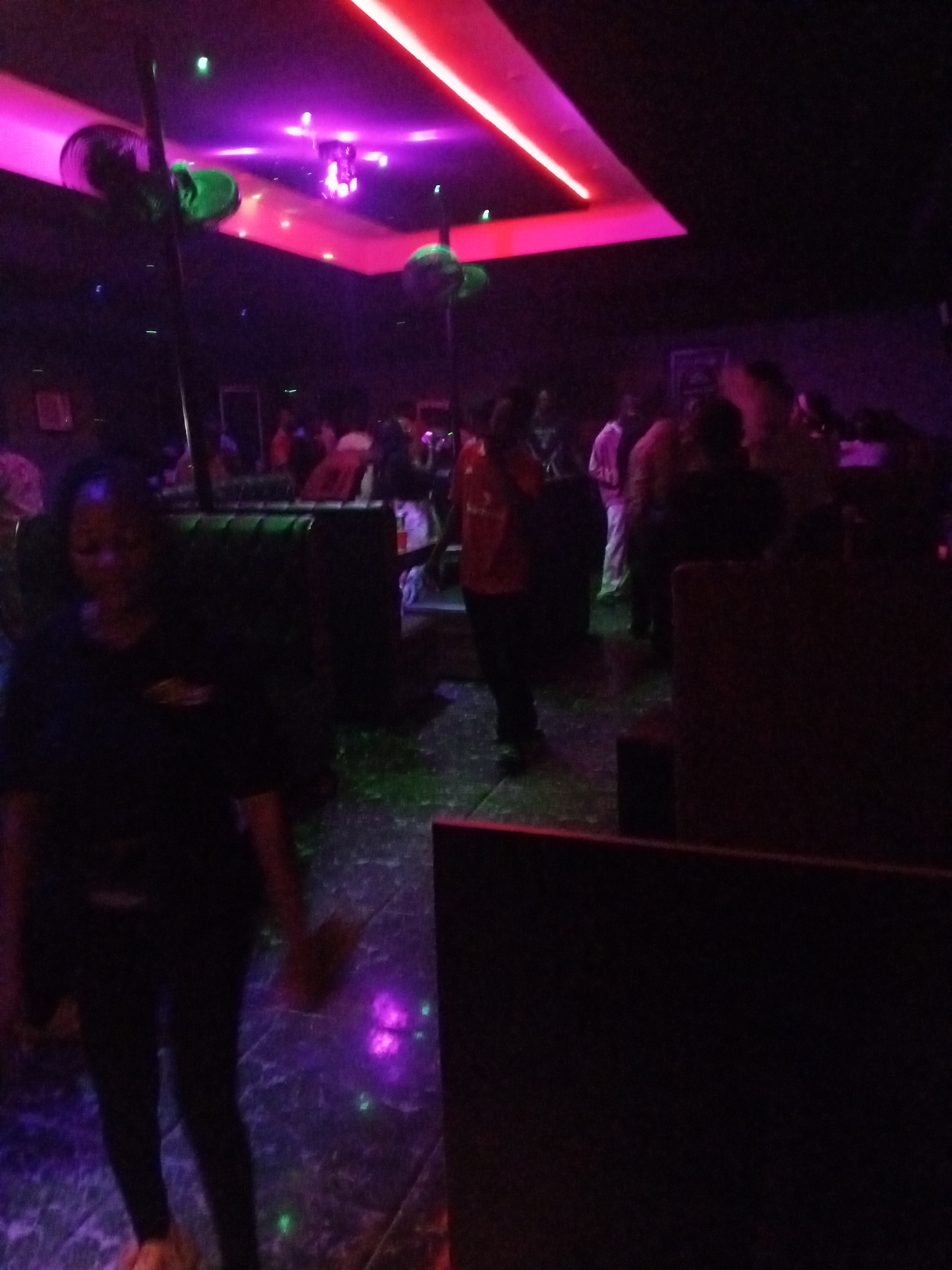The image shows a dimly lit interior, likely a nightclub or bar, located in Lokoja, Nigeria. The lighting features pink and purple hues, with green spotlights on the ceiling. Several people are present, engaged in conversations or moving around. A woman is in the foreground, wearing dark clothing. There are tables and seating areas, as well as reflective flooring with light patterns. Some objects include the ceiling fan. The environment is dark and energetic, typical of a nighttime entertainment venue.