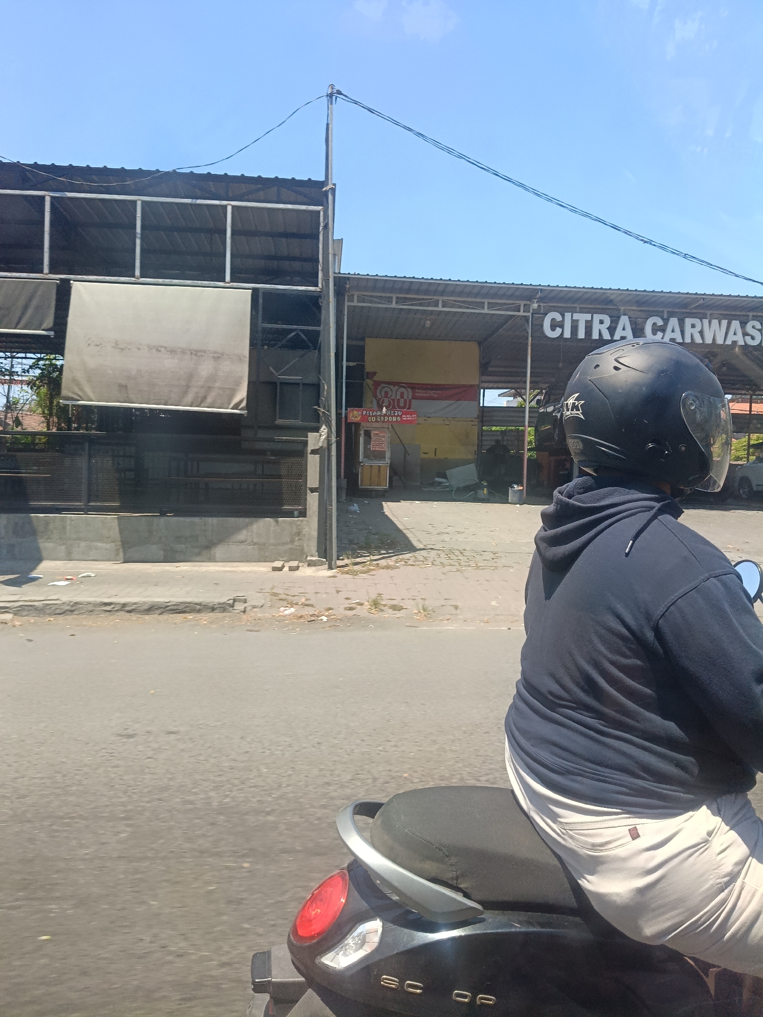 In Buduran, Indonesia, under a clear blue sky, a person wearing a navy blue hooded sweatshirt and light-colored pants rides a black scooter. They are also wearing a black helmet. The scooter is driving on a paved road, passing by a car wash establishment. The car wash has a sign that reads "CITRA CARWASH." The building appears to be an open-air structure with a corrugated metal roof supported by metal poles. There are also some trees and other buildings visible in the background. The area appears to be a commercial district.
