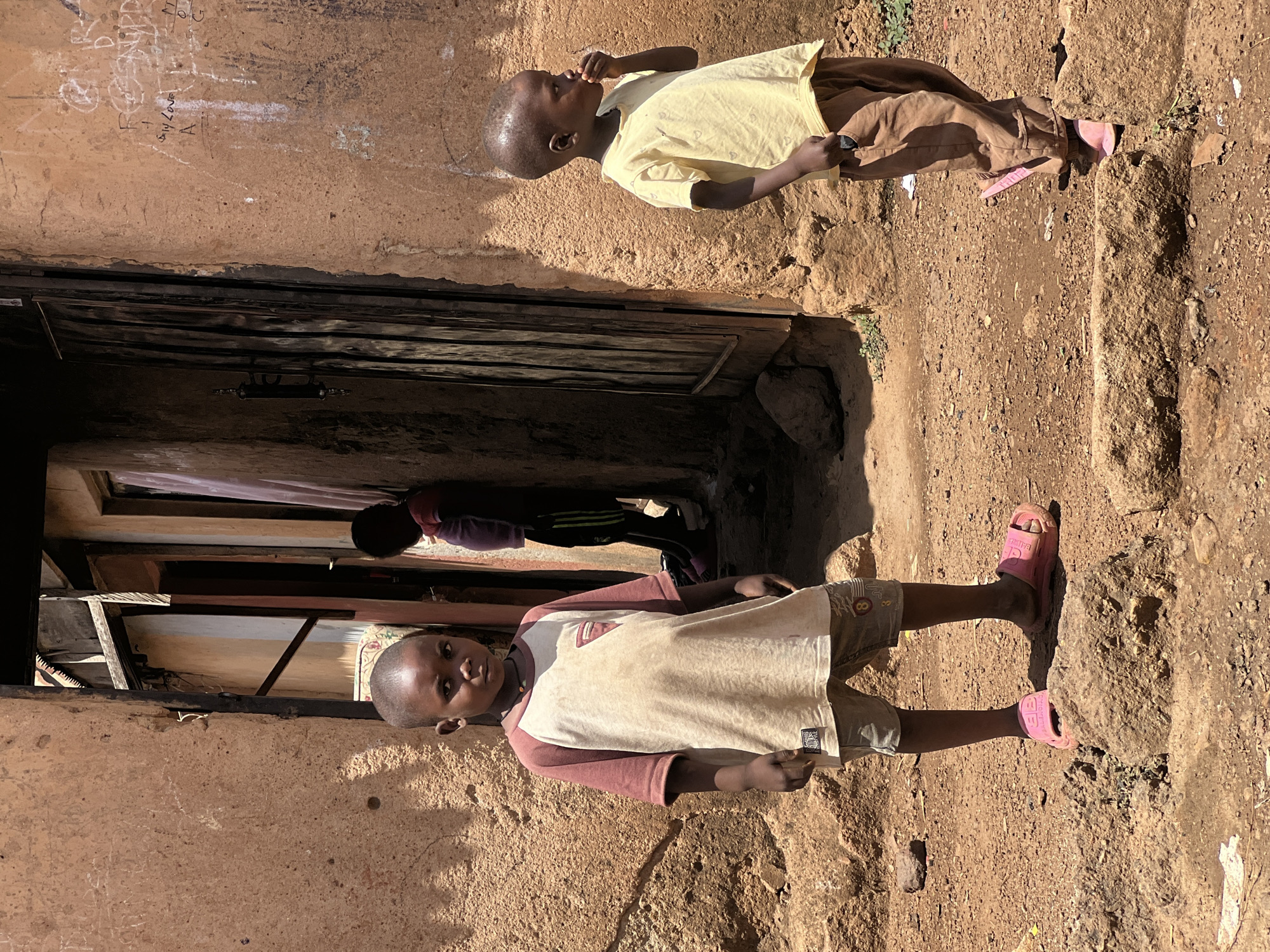 Two African children stand in front of a building in Jos, Nigeria. The building has a dark doorway. One child wears a light-colored shirt and brown pants, and the other wears a cream-colored shirt with maroon sleeves and brown shorts. Both children are wearing pink sandals. The ground is dirt.