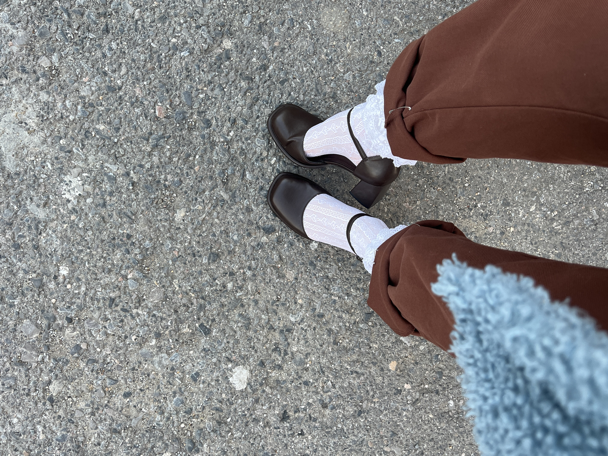 The image is a downward-facing shot of a person standing on asphalt. The person is wearing brown trousers cuffed at the ankle and white lace socks paired with dark brown Mary Jane shoes with block heels. Part of a light blue, fluffy item is visible in the lower right corner of the frame. The location cannot be determined as Palilula Urban Municipality, Serbia, from the provided image.