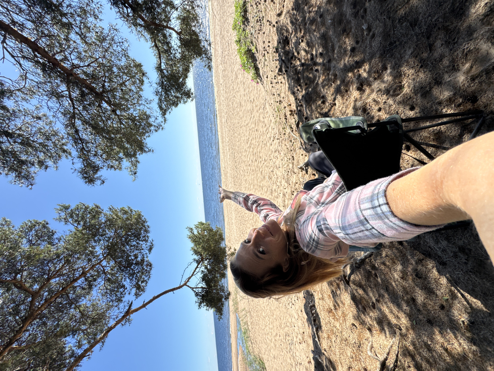 A woman is pictured from a first-person perspective on a sandy beach, with pine trees and the sea in the background. She is wearing a pink and gray plaid shirt and pointing with her left hand towards the sea. A foldable camping chair is visible behind her, with a green bag on it. The sun is shining, casting shadows from the trees on the sand, suggesting it is daytime. The water appears calm and the sky is clear blue. The scene evokes a sense of leisure and relaxation in a natural outdoor setting, possibly a vacation or day trip. No visible text is present in the image.