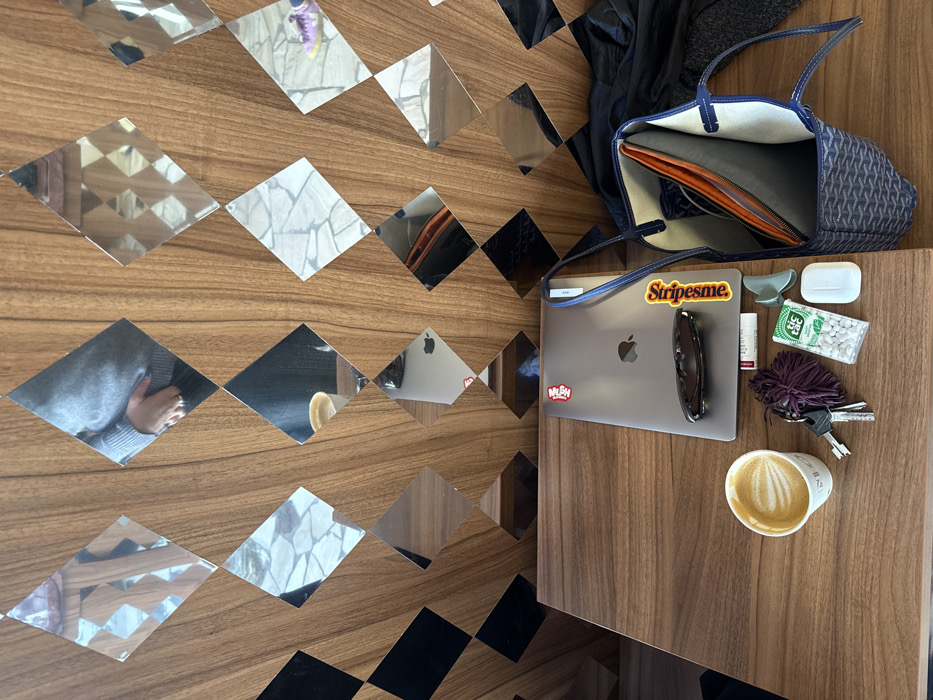 The scene is set on a dark wooden table within a cafe located at 139 Rue Saint-Honoré in the 1st arrondissement of Paris, France. The cafe's wall features wooden paneling with vertical striations, accented by a repeating pattern of diamond-shaped mirrors and black diamond inlays. Mirror reflections show a person wearing a light gray long-sleeved top, with their right hand visible near their torso, along with sections of a patterned stone floor and a purple sneaker.
On the dark wooden table, from left to right, are: a white disposable coffee cup containing a latte with a fern-like art design; a set of silver keys attached to a dark purple fluffy keychain; a closed silver Apple MacBook laptop adorned with "Nulogy" and "Stripes.me" stickers, upon which a pair of dark-tinted sunglasses rests; a small white rectangular item; a green container of white Tic Tac mints; a white Apple AirPods charging case; and an open blue Goyard tote bag with its signature chevron pattern, revealing a light interior lining and an orange pouch. A dark-colored garment is draped to the right of the tote bag, partially on the table. No direct actions or interactions are depicted beyond the arrangement of items and the reflected partial view of a person.