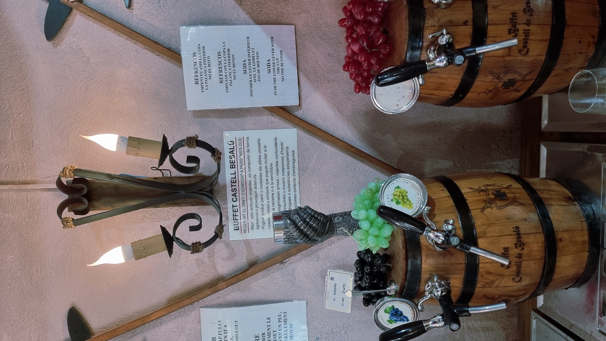 The image shows a close-up view of a buffet setup in Besalú, Spain, featuring decorative elements and beverage dispensers. In the background, a rustic wall sconce with two lit candle-shaped bulbs hangs above a sign for "BUFFET CASTELL BESALÚ" in Catalan. This sign details rules for admission, including prohibitions against violent behavior, aggression, and disruptive actions. To the right, two separate signs provide instructions in Catalan and French for operating a beverage dispenser, referring to "REFRESCOS" (soft drinks) and "SODA."
In the foreground, two wooden barrels, designed to resemble wine casks, serve as bases for beverage taps. Each barrel has a decorative crest featuring crossed swords and a shield, along with the inscription "Buffet Castell de Besalú." The left barrel is adorned with a cluster of green grapes and a small sign indicating "VI NEGRE" (black wine) and "VINO TINTO" (red wine). Above the green grapes, a circular emblem features a cluster of dark grapes. The right barrel is decorated with a cluster of red grapes. Both barrels are equipped with chrome beverage taps. The scene is set in what appears to be an indoor dining area, with a metal serving counter visible at the bottom. The lighting is dim, with the candles in the sconce providing a warm glow.