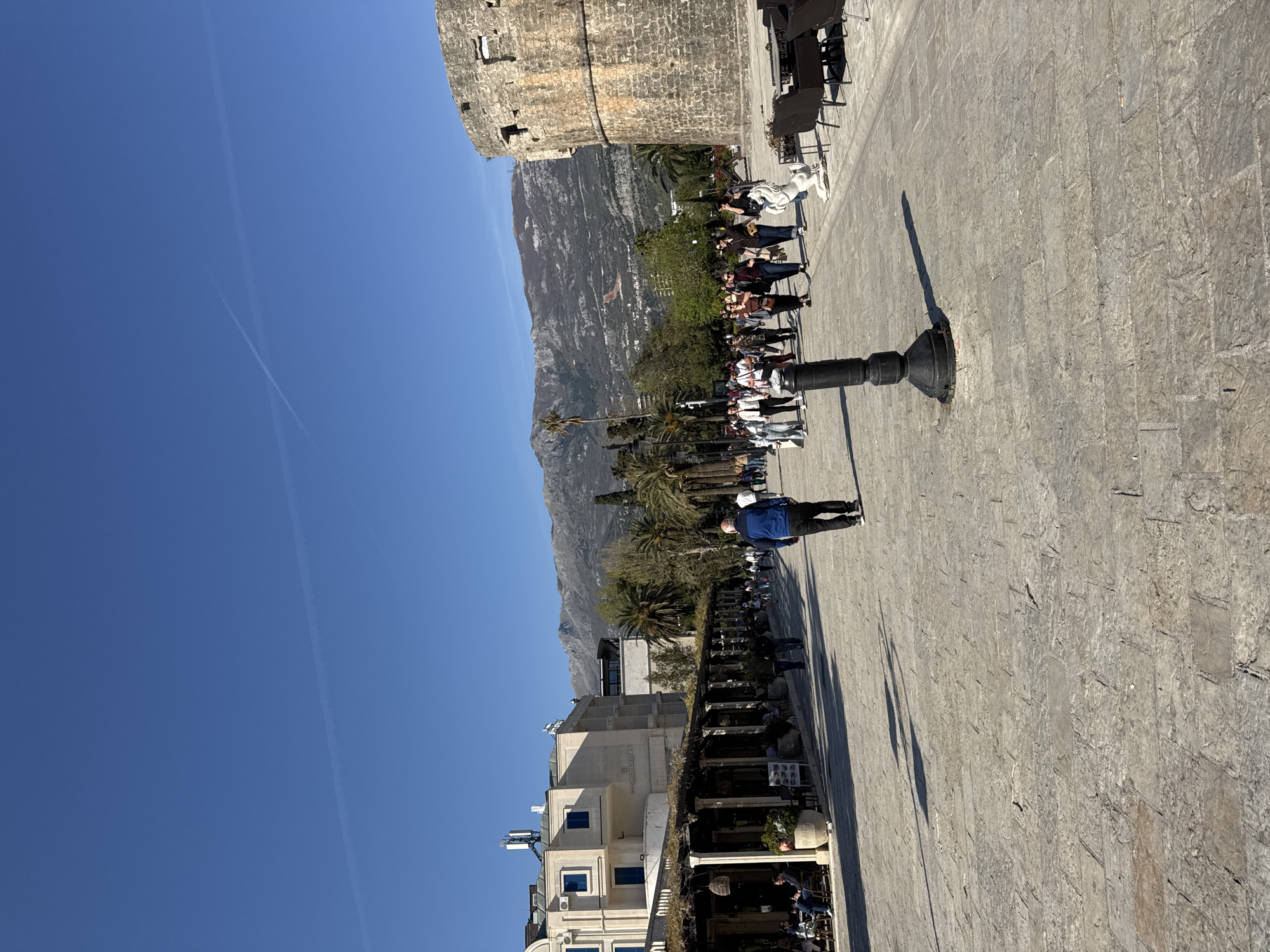 The image shows a paved public square in Budva, Montenegro, possibly near the old town fortifications. The sky is clear blue with contrails. A stone tower structure is visible in the background to the left. People are sitting at outdoor tables, likely part of a restaurant or café along the right side of the frame. The terrain slopes upwards towards mountains in the mid-ground. A single individual stands in the middle of the square. A dark-colored, short, cylindrical object, possibly a bollard or the base of a light fixture, stands on the paving. Buildings with white facades are seen on the left.