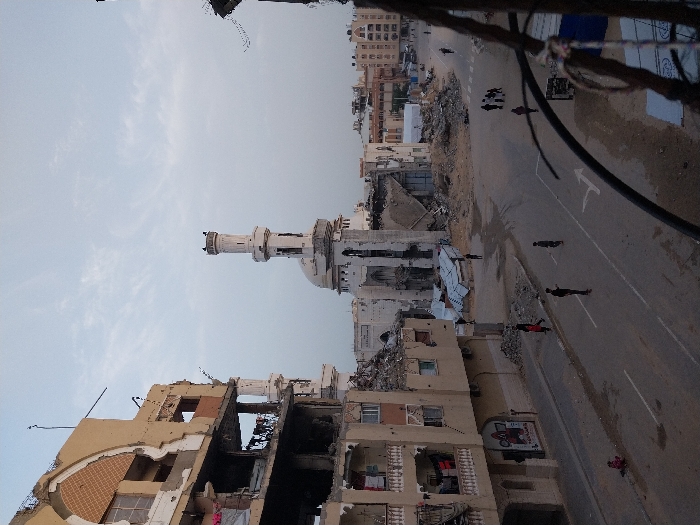 A damaged mosque with a minaret stands amidst destroyed buildings, rubble, and a street. Several individuals are walking along the street, which has a painted traffic arrow. Other intact buildings are in the background. The image shows destruction and potential human movement in a post-conflict urban environment.