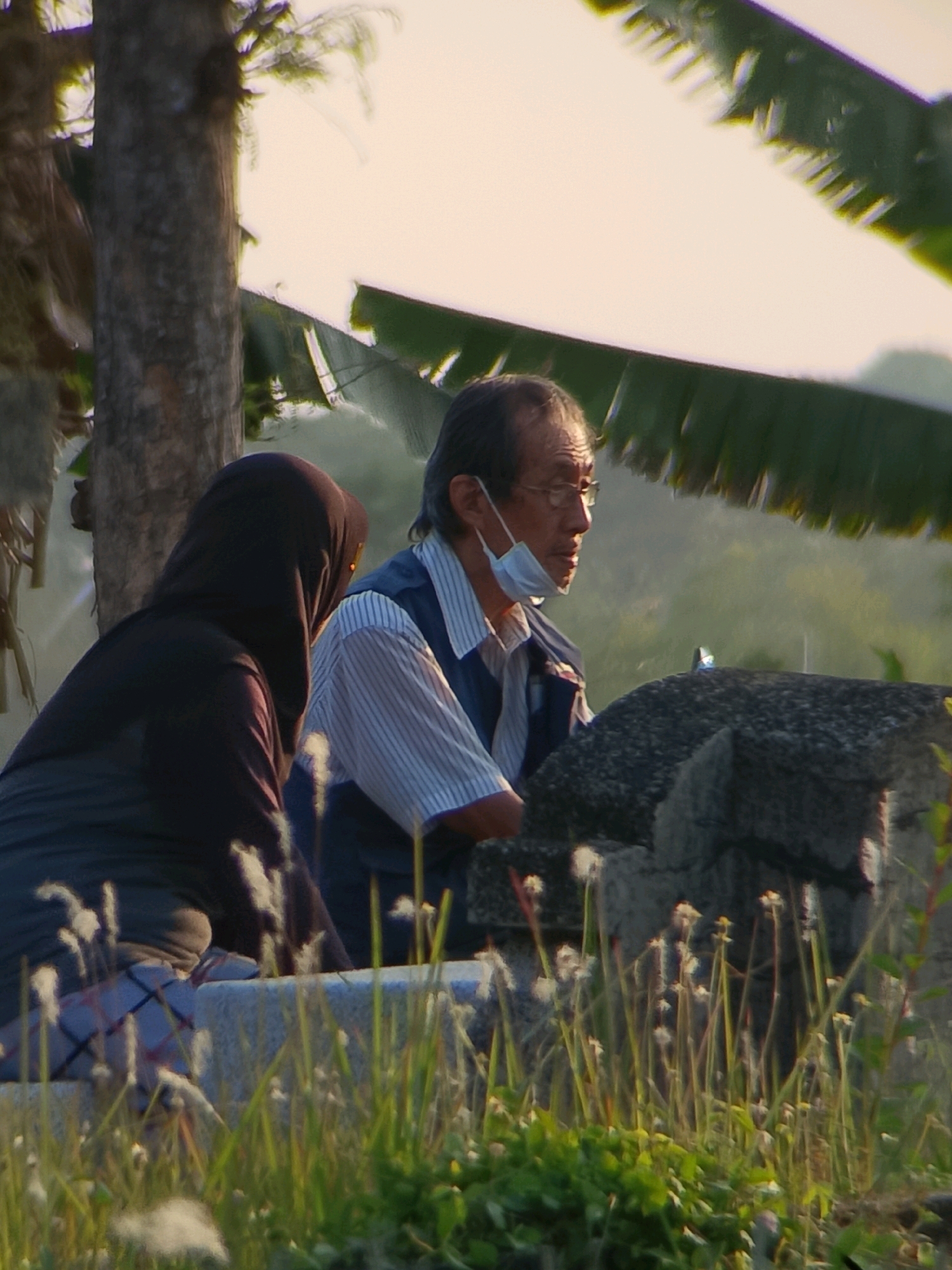 The image depicts two individuals outdoors in Jember, Indonesia. On the left, a person is wearing a dark hijab and a dark top, with a checkered pattern visible on their lower garment. They are seated on a low stone wall. To their right, an older man with glasses is wearing a blue vest over a striped shirt and has a face mask pulled down below his nose. He is also seated on the stone wall, facing towards the left of the frame and appears to be looking downwards. The background features a tree trunk on the left, large green leaves from a plant overhead, and a blurred, hazy backdrop suggesting a natural or semi-rural environment. Tall grass and small white wildflowers are in the foreground, growing around the stone wall. The lighting indicates it is either early morning or late afternoon.