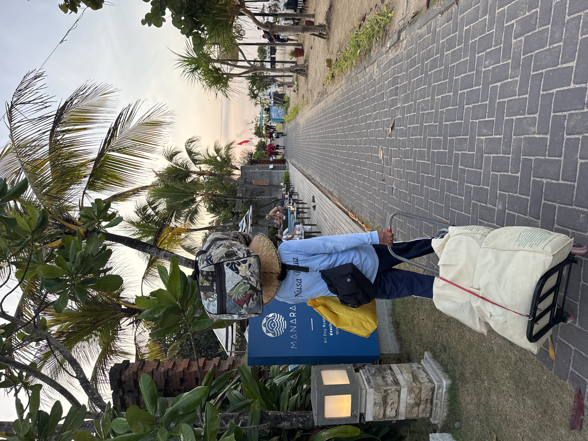 A person wearing a blue long-sleeved shirt, dark pants, and a straw hat is walking on a brick pathway. The hat has two bags stacked on top of it. A yellow bag is slung over the person's right shoulder, and a black bag is hanging from their left shoulder. The person is pulling a luggage cart with two large, light-colored sacks. Behind the person, a blue sign reads "MANARA" with the text "Nusa Dua" below it, and "All Day Restaurant Beach Bar Wine." The pathway is lined with palm trees and greenery. In the background, there are outdoor seating areas with tables and chairs, some people, and the ocean. The setting appears to be a beachside resort or restaurant in Nusa Dua, Kuta Selatan, Indonesia, during the late afternoon or early evening, indicated by the soft lighting and the direction of the sun.