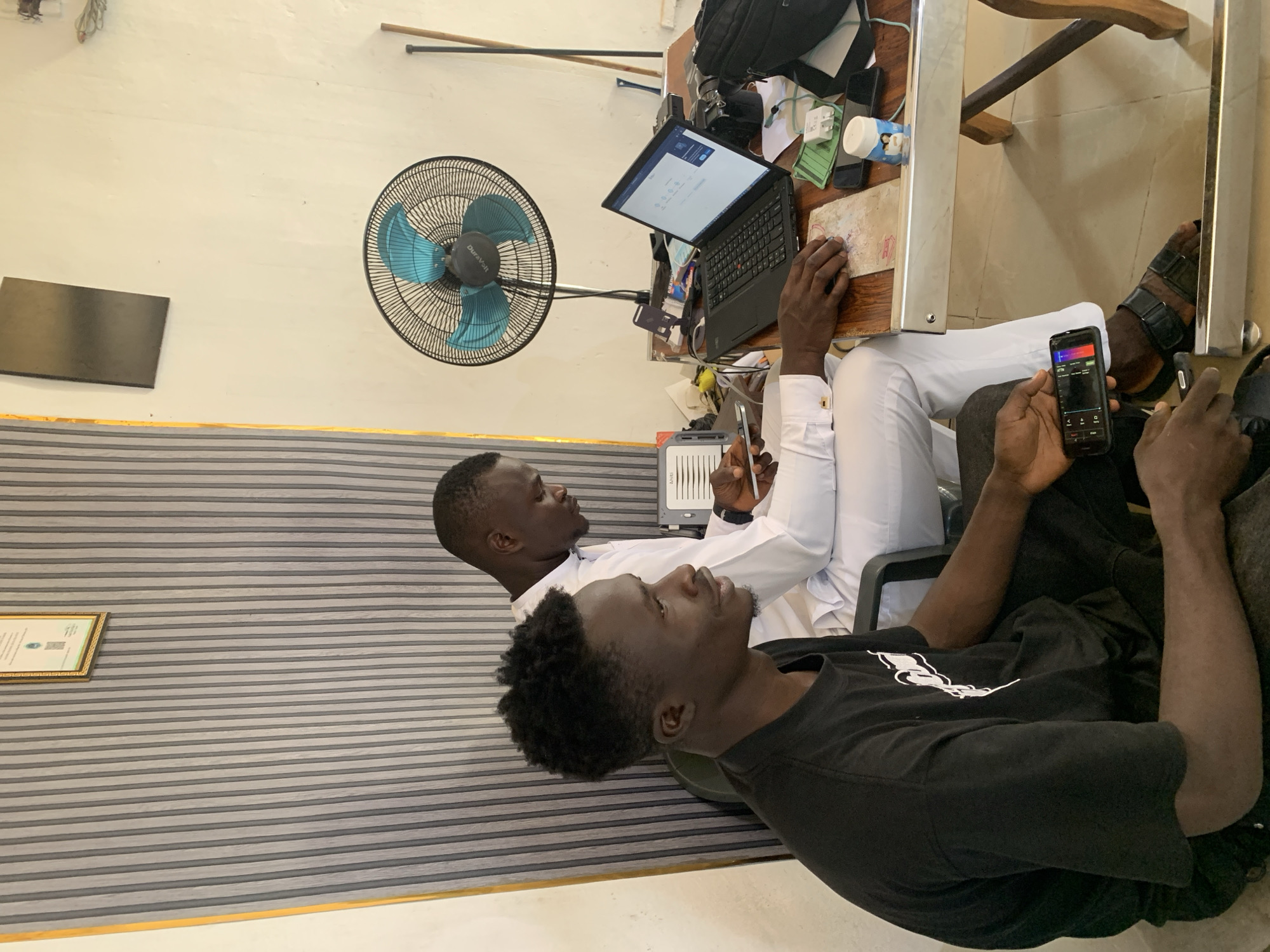 In a room in Jos, Nigeria, two men are seated at a wooden table. The man on the left, wearing a black t-shirt, is looking to his right while holding a smartphone in his left hand. The man on the right, dressed in white, is facing the table and typing on a laptop. The table is cluttered with various items, including a fan, a laptop, a phone, and a small container with an image of a child. The room's walls are painted white, and one wall is partially covered by a striped curtain.
