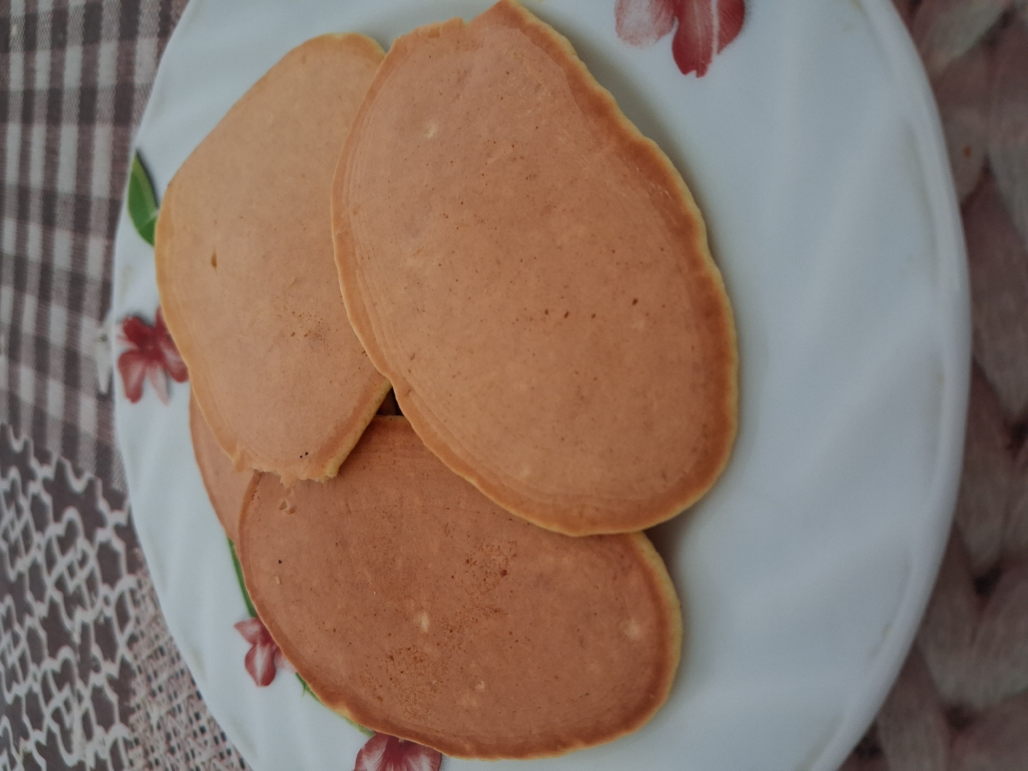 A close-up shot shows three golden-brown pancakes arranged on a white plate with a floral pattern. The pancakes are slightly overlapping, with the largest one in the foreground. The plate rests on a patterned tablecloth with brown and beige squares and a floral motif. The background is out of focus. There are no people visible in the image. The scene appears to be indoors, possibly in a home setting in Krolevets, Ukraine.